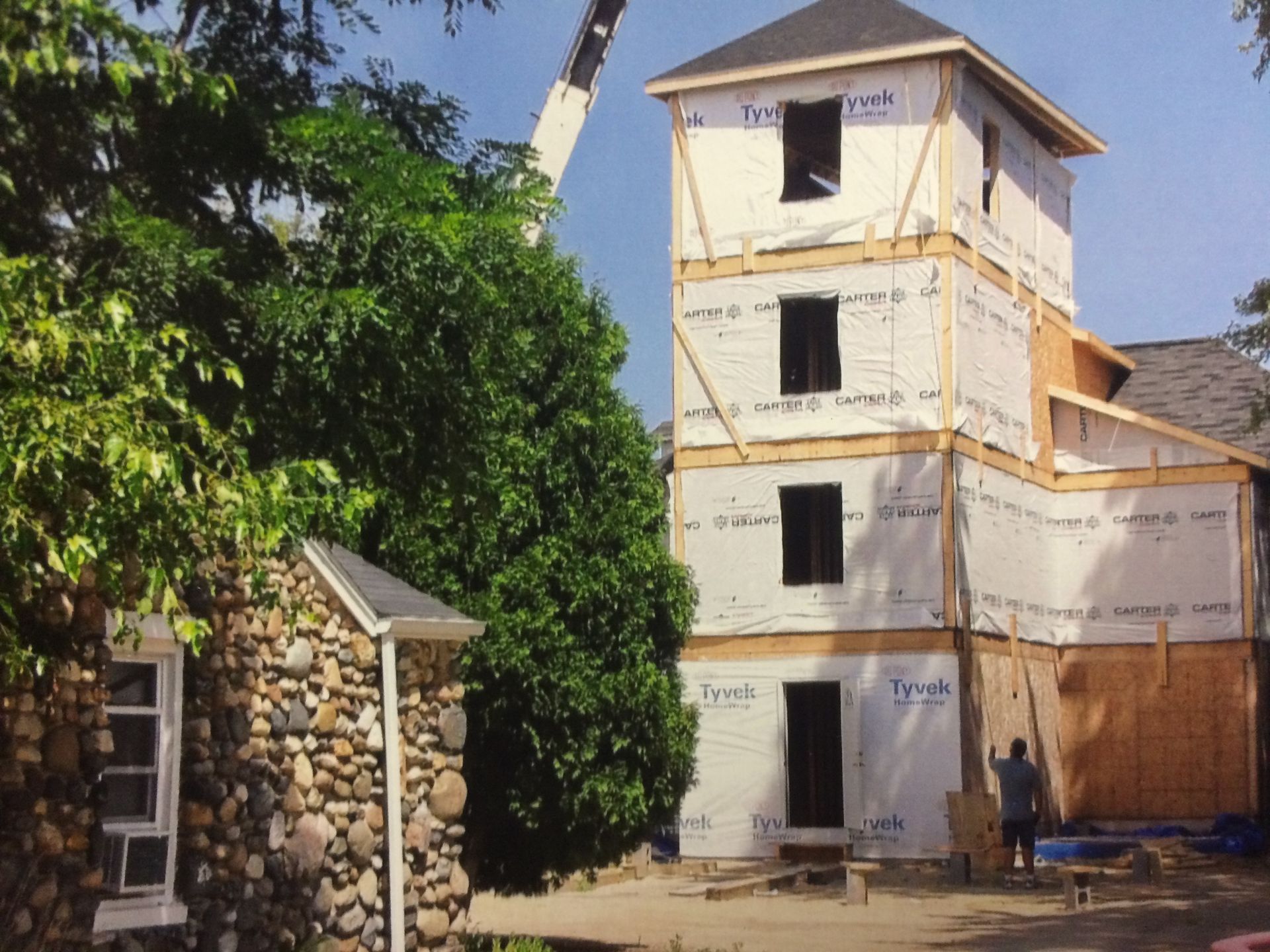 Building under construction; a three-story tower with Tyvek wrap and a crane in the background next to a stone building.