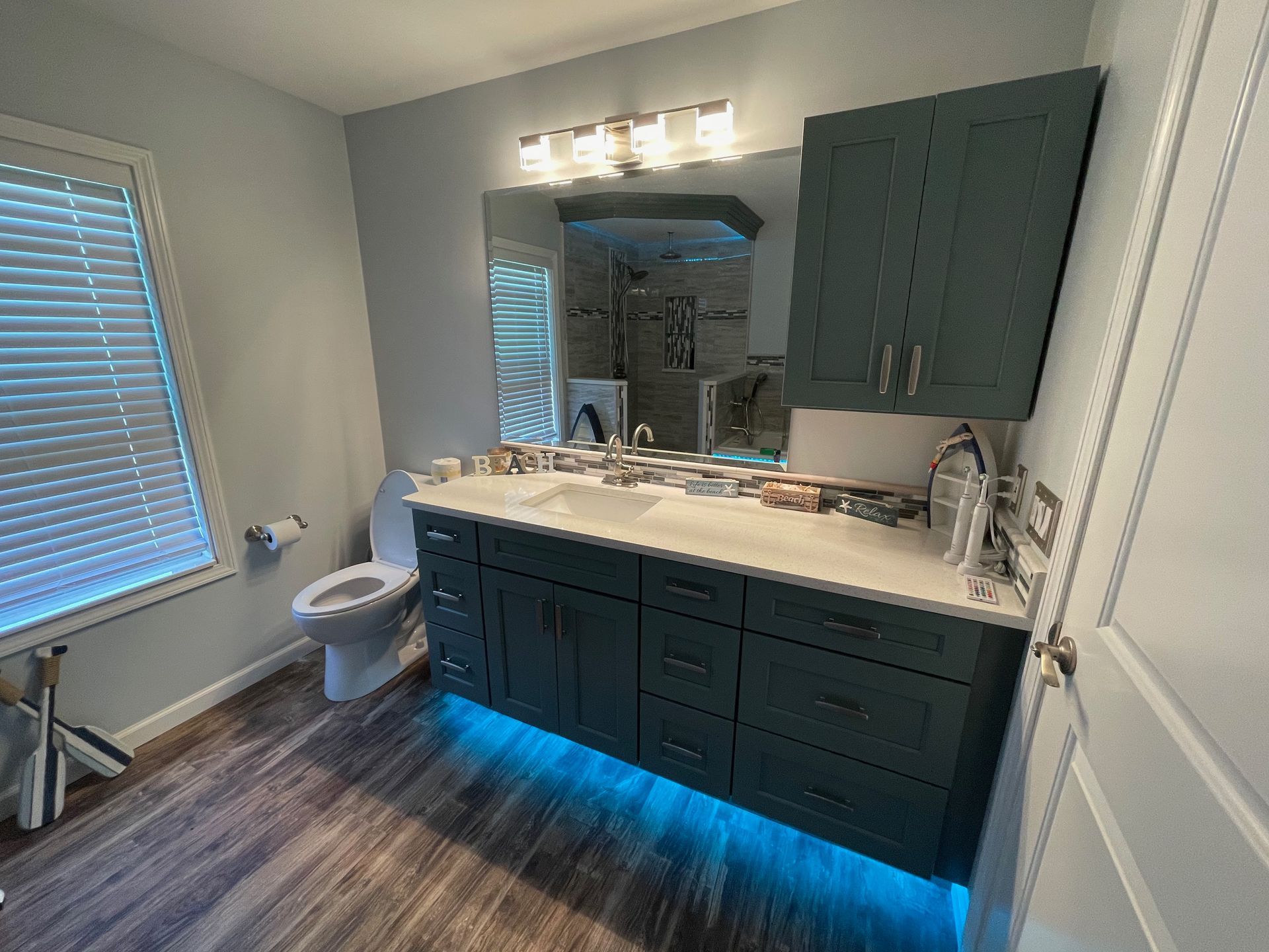 Bathroom with teal cabinets, wood-look floor, large mirror, and under-cabinet LED lights.
