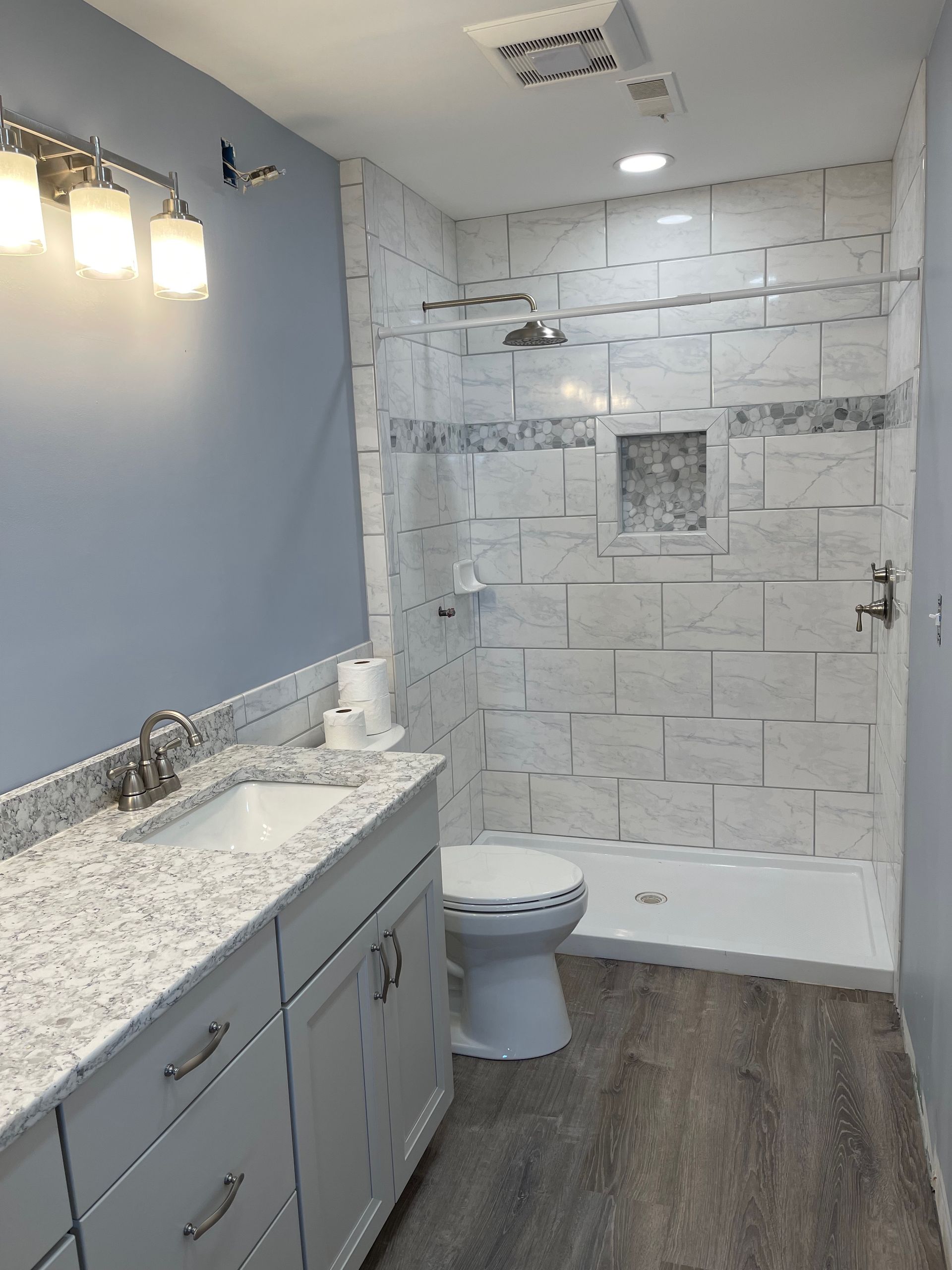 Bathroom with gray cabinets, a white sink, and a tiled shower. Light blue walls and gray flooring.