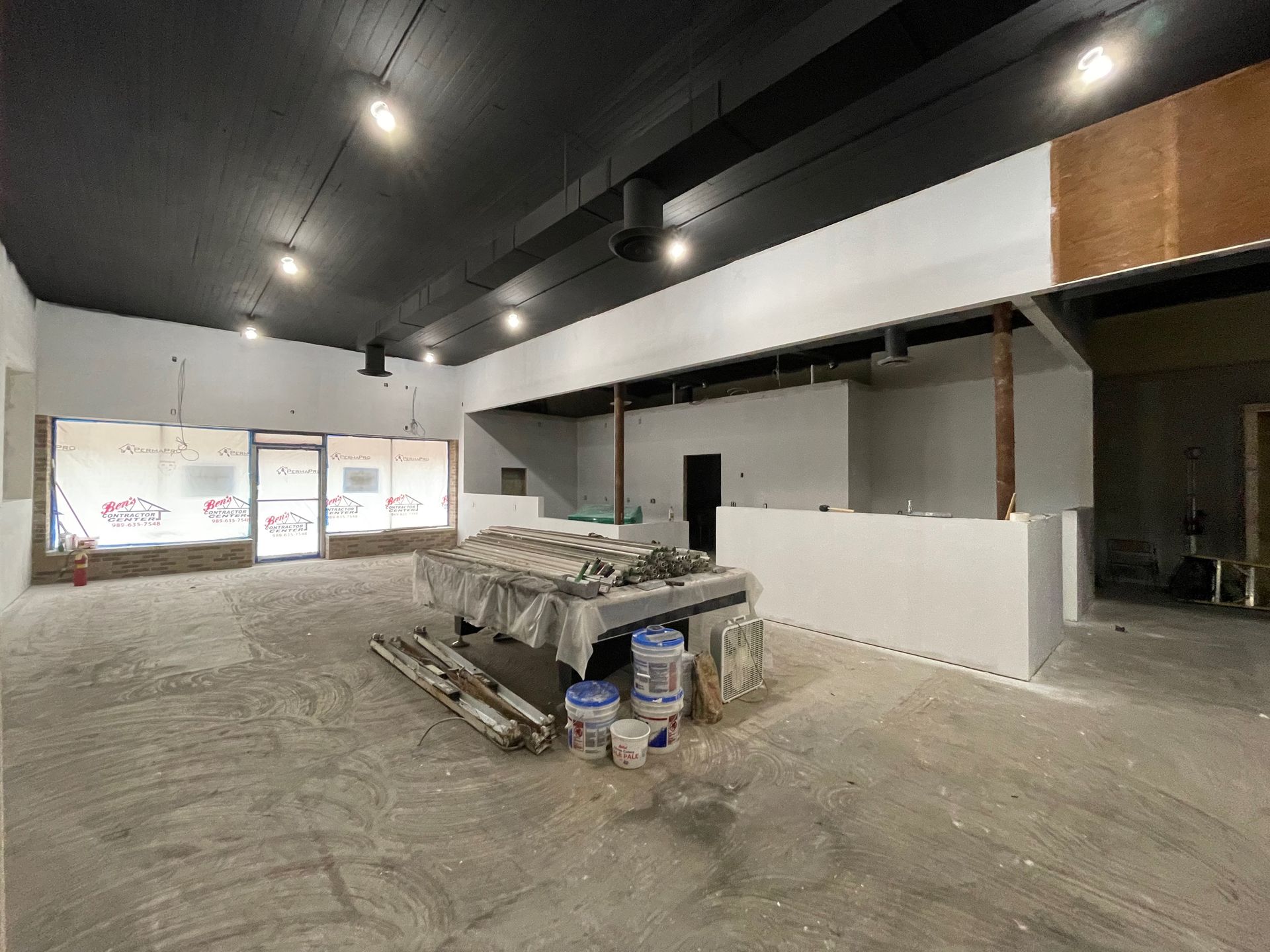 Interior of a space under construction; white walls, black ceiling, construction materials in the center, and a storefront entrance.