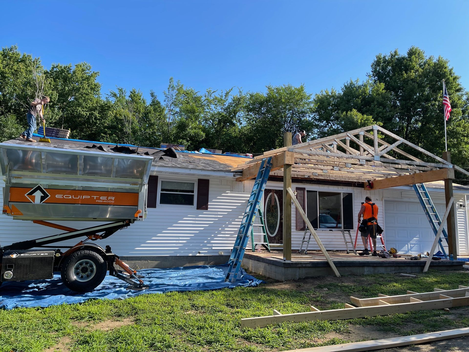 Roof being replaced on a white house with a new porch under construction; a debris trailer is beside it.