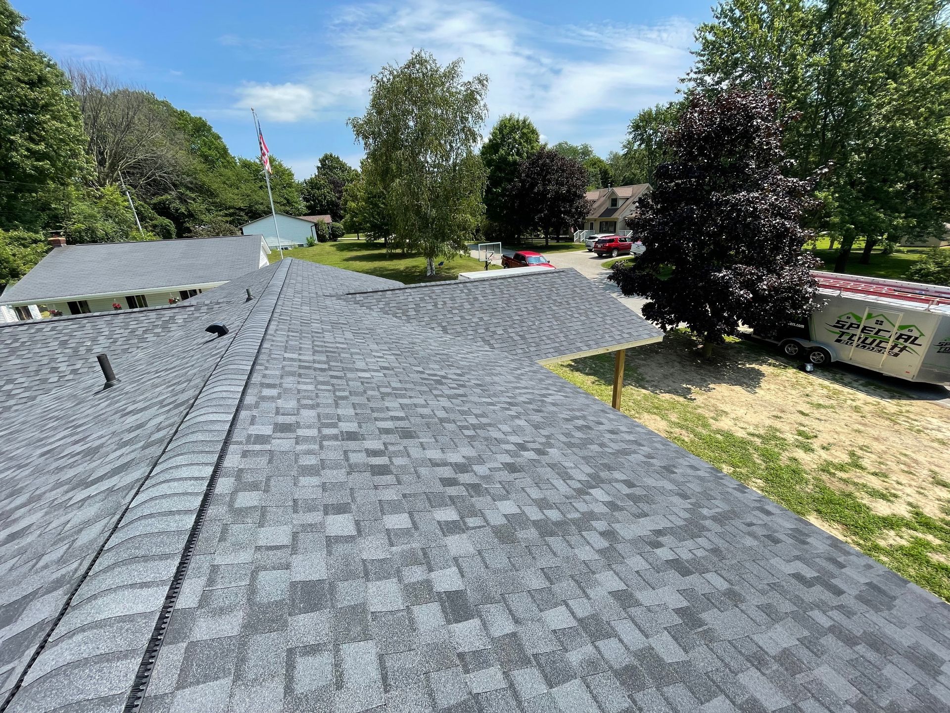 Gray asphalt shingle roof on a house, under a sunny sky. Trees and a red truck are in the background.