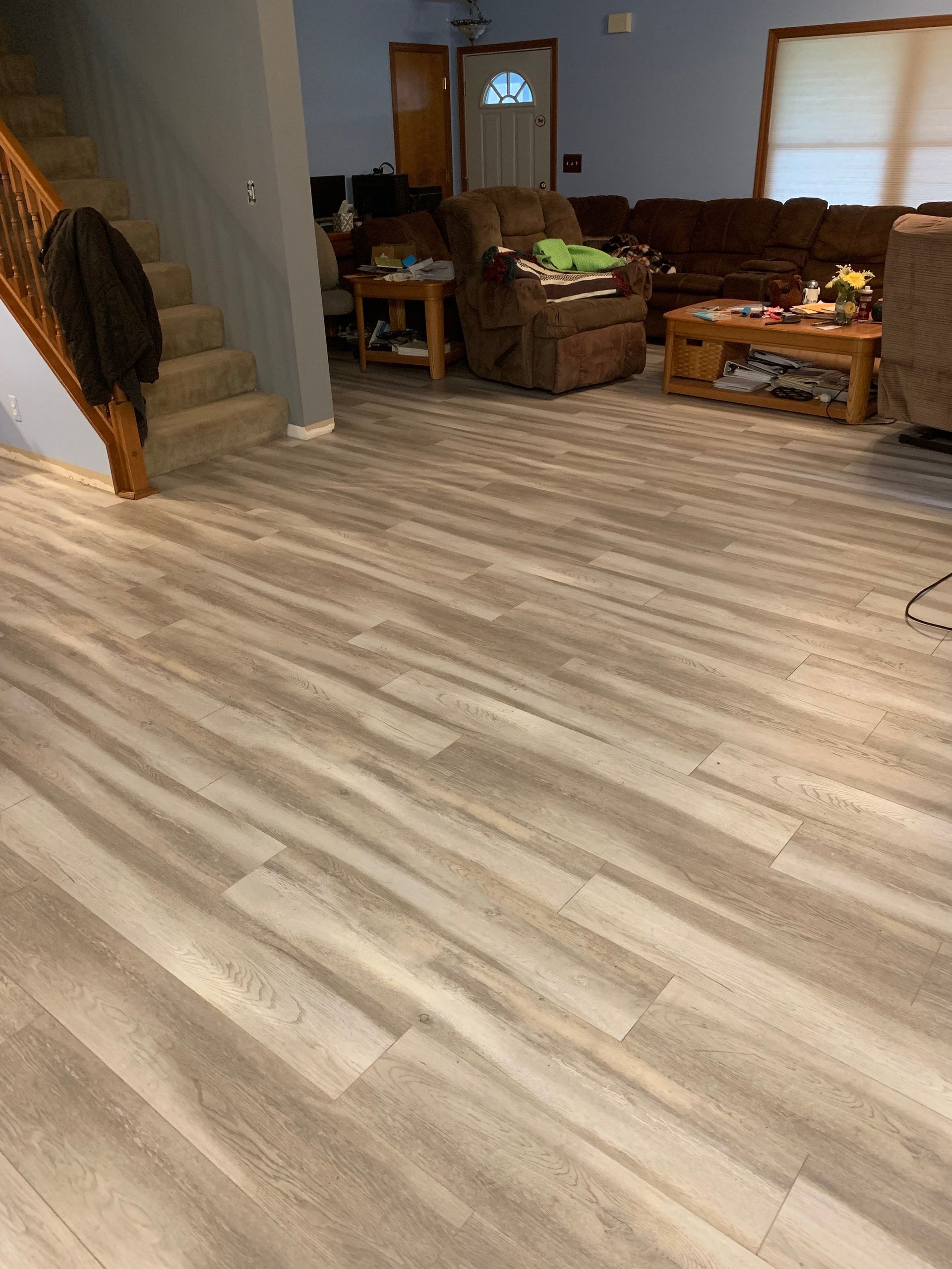 Living room with wood-look flooring. Brown recliner and sofa, wooden coffee table, and stairs leading up.