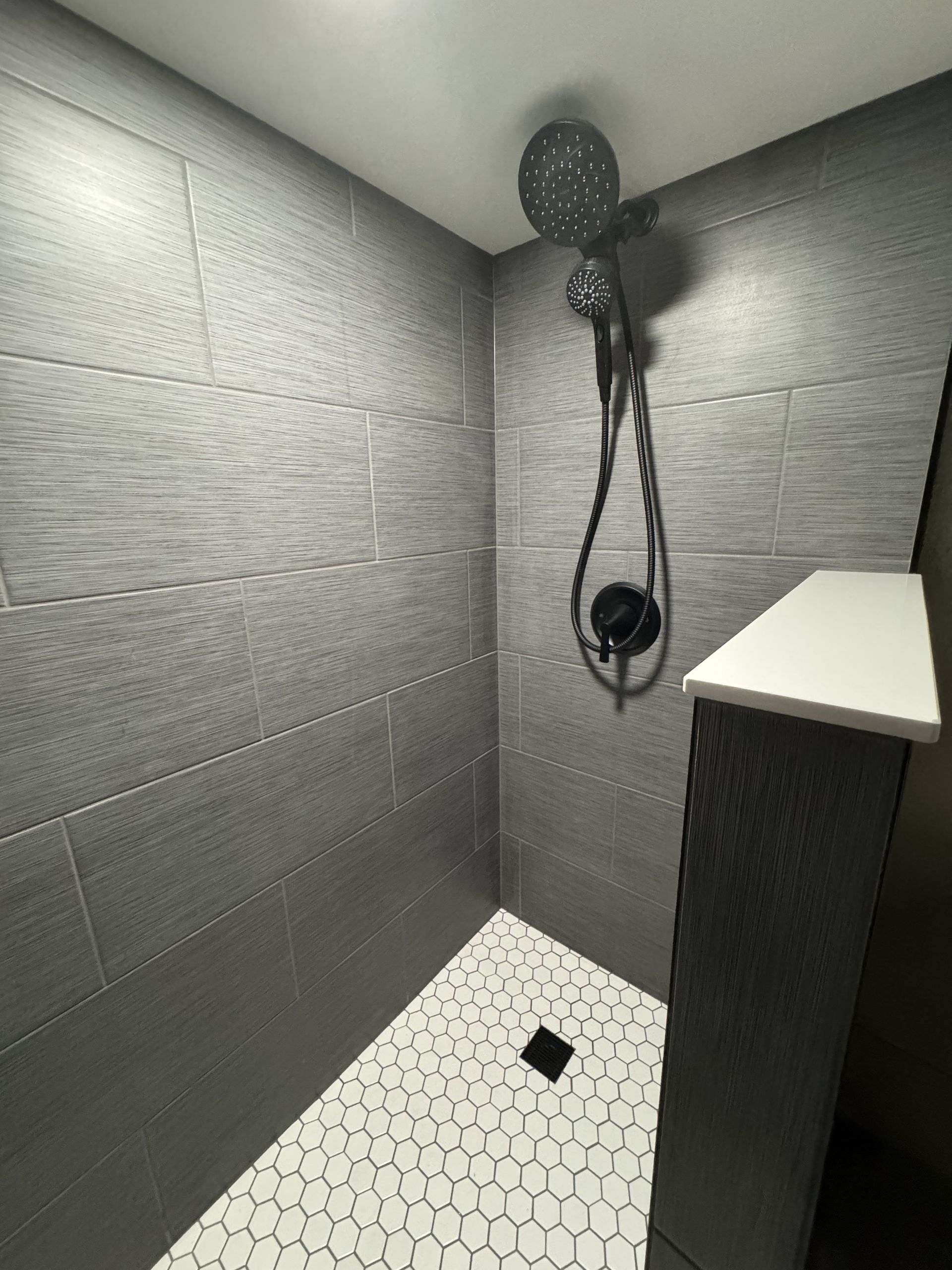 Gray-tiled shower with pebble-tile floor, black shower fixtures, and a small built-in shelf.