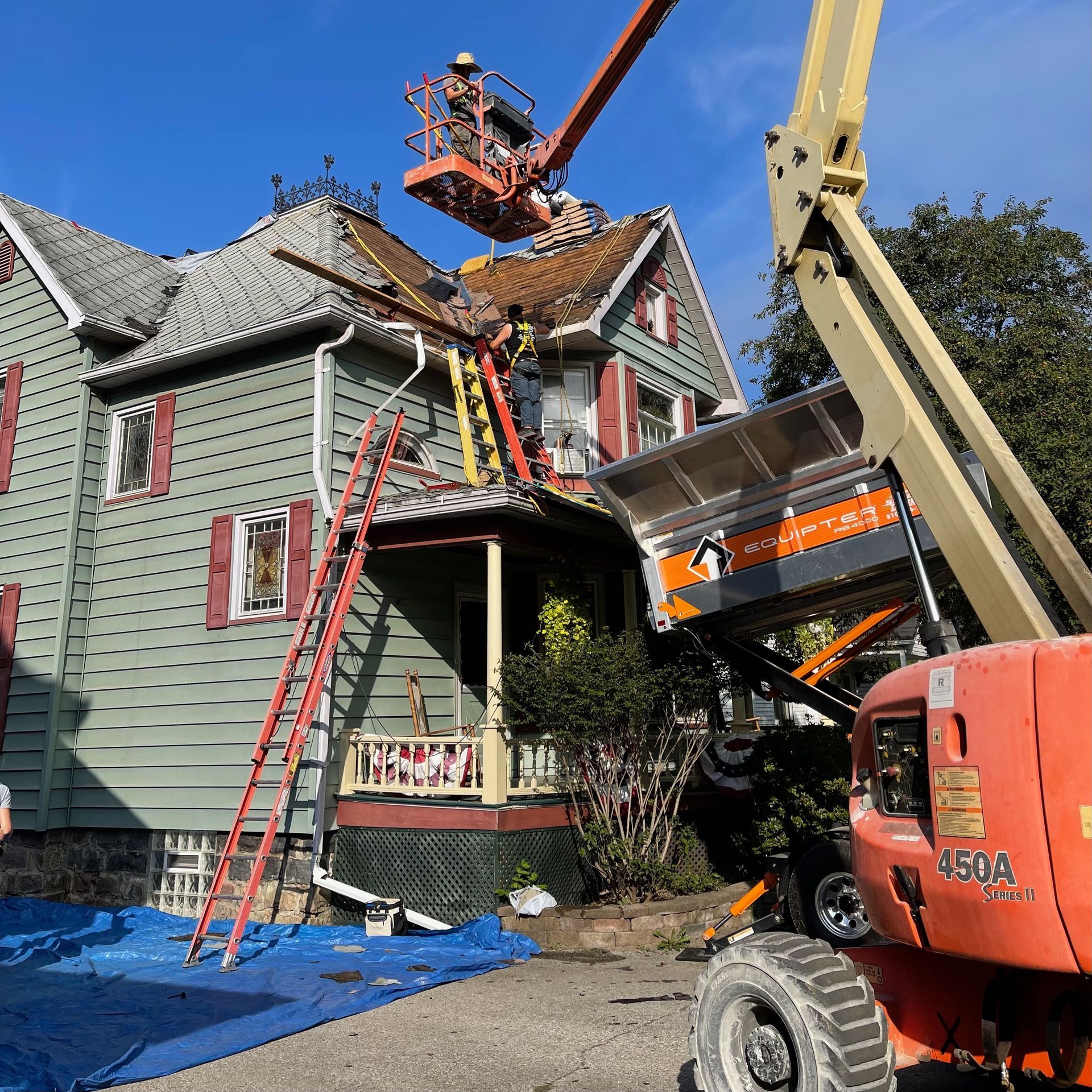 Workers on lift repairing roof of a two-story green house with red shutters, orange and black lift in front.