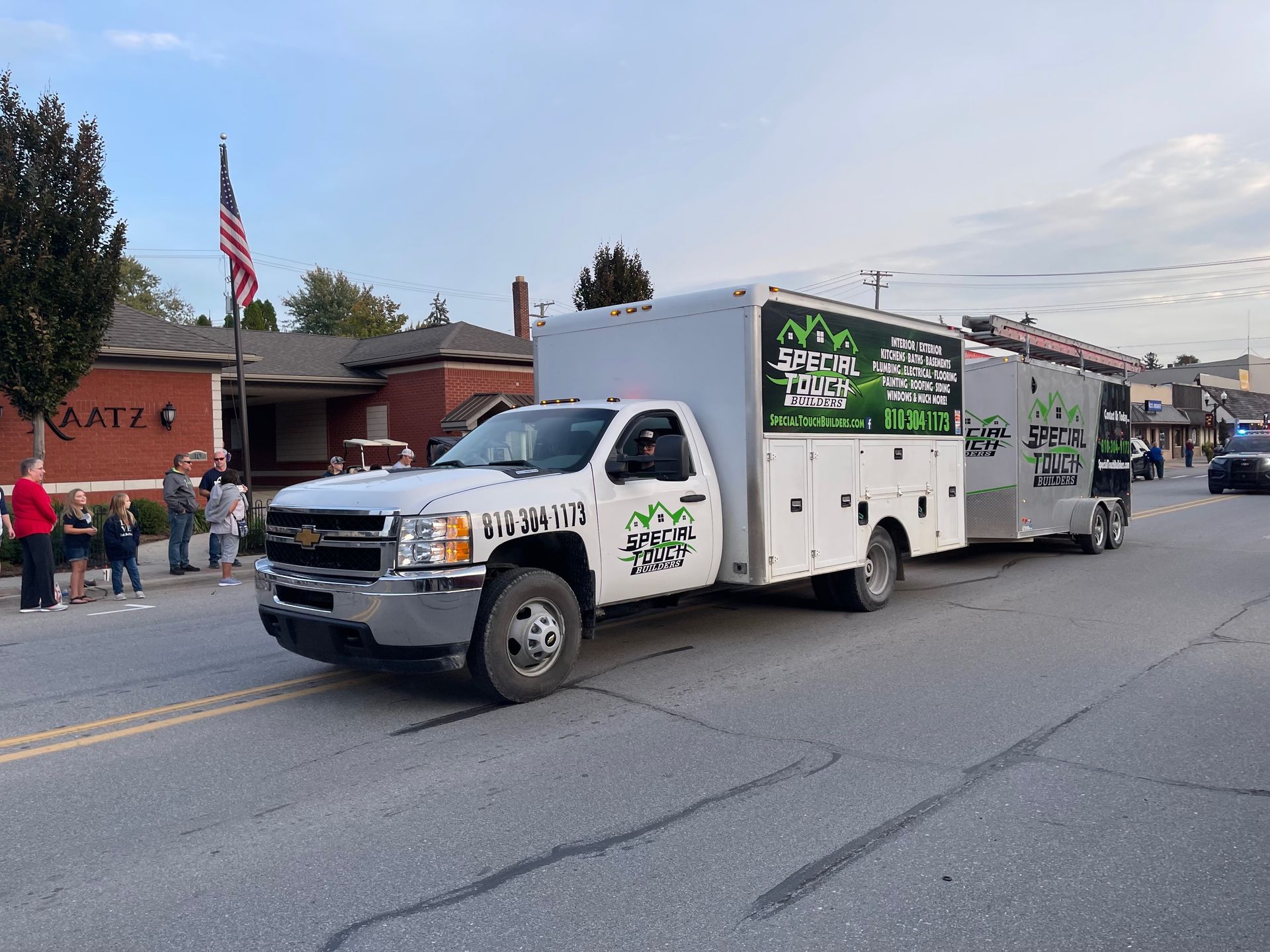 A white work truck towing a trailer with company logos drives down a street during a parade; people watch.