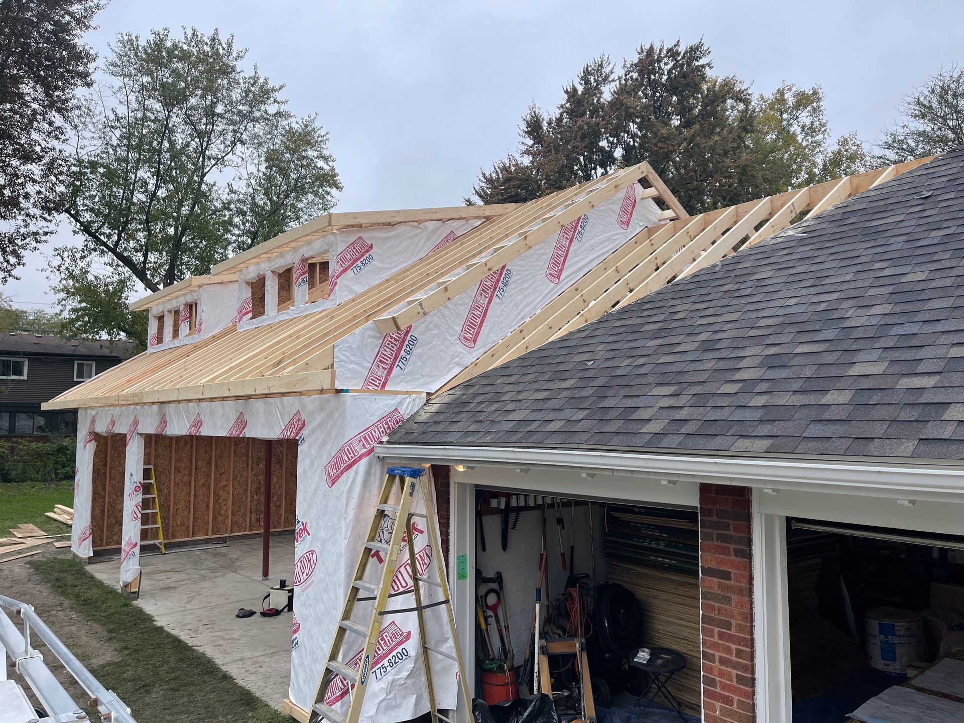 Garage construction in progress; wooden frame, partially covered with wrap, adjacent to existing garage.