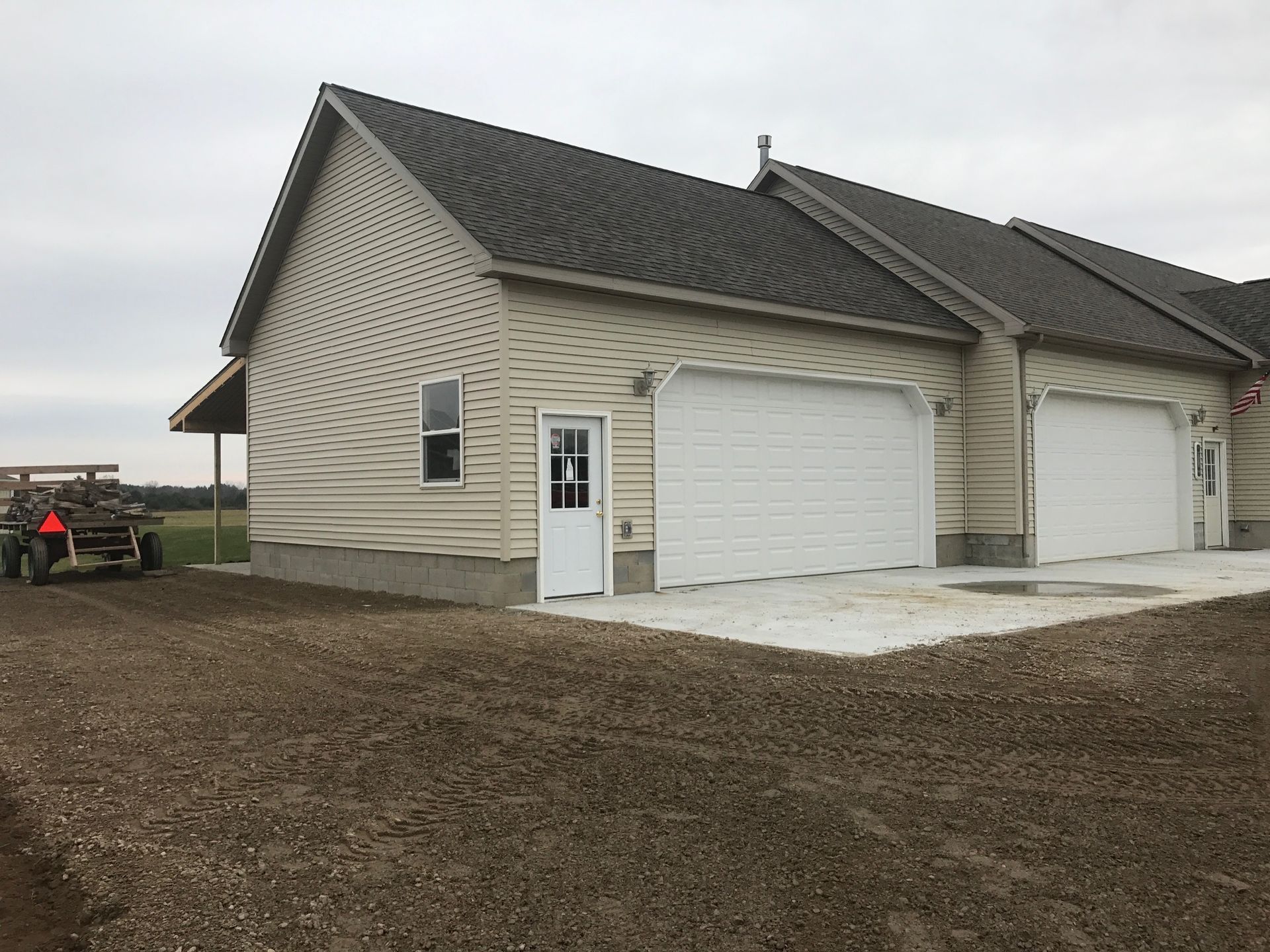 Light beige garage with white doors, gravel driveway, and a tractor in the distance. Cloudy sky.