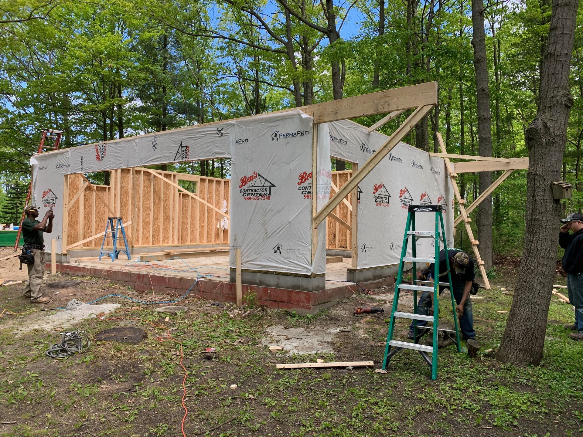 Construction of a wooden building in a wooded area; workers install boards, wrapped with waterproof material.