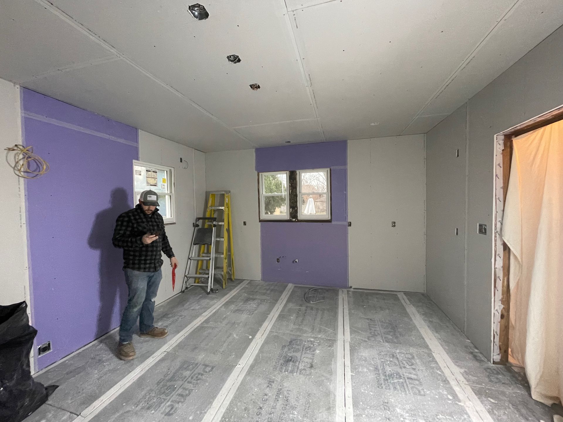 Man in room under construction; purple and grey drywall, white floor lines, open doorway, ladder.