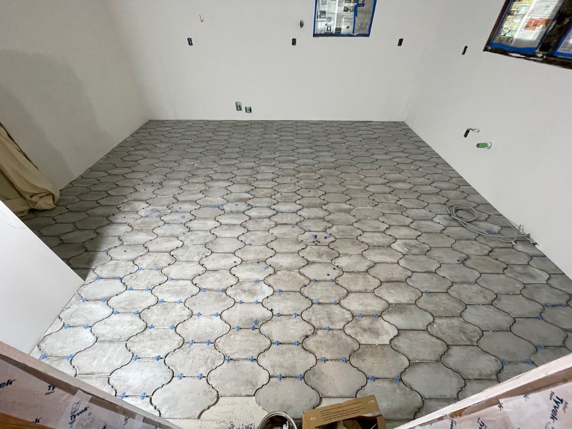 Room with gray hexagonal tiles being laid on the floor, unfinished walls, bright overhead lighting.