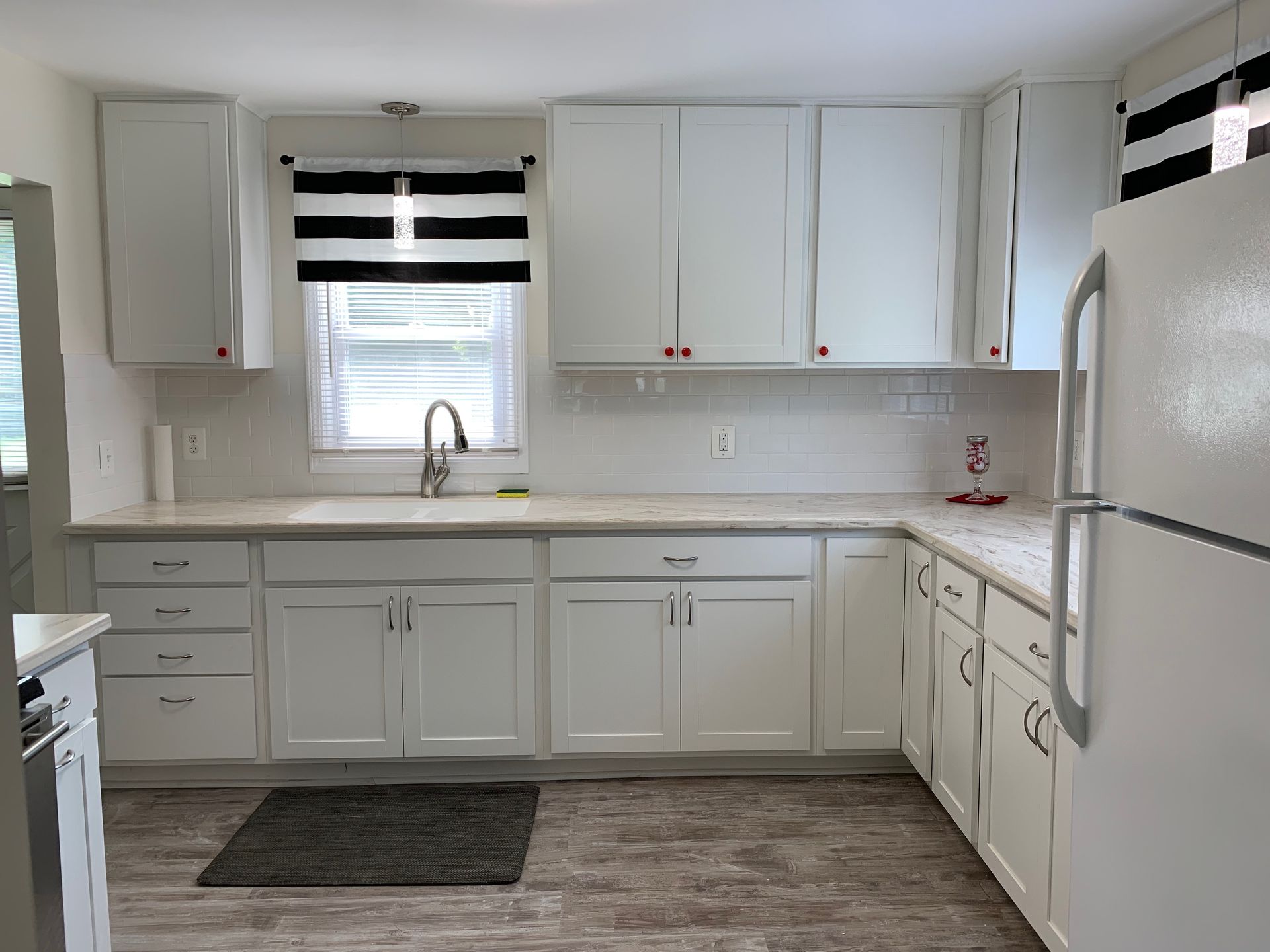 White kitchen with cabinets, countertops, window, and refrigerator. Gray rug on floor.