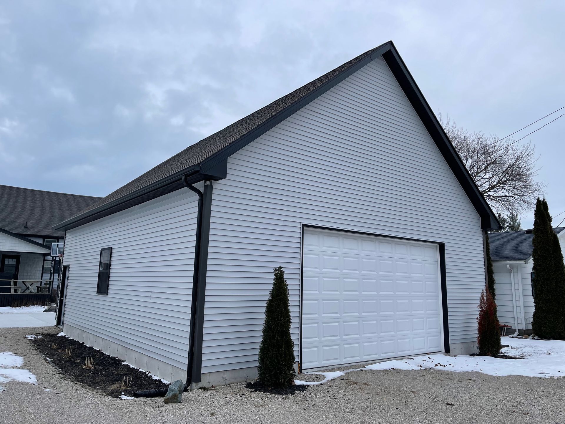 White-sided detached garage with a white garage door, black trim, and a gray roof on a gravel driveway.