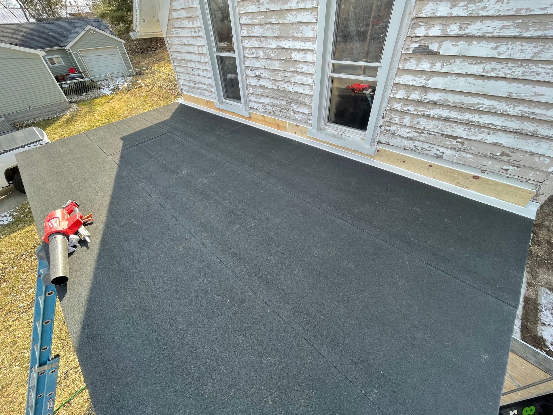 View of a flat roof with new black roofing material next to a white house with windows.