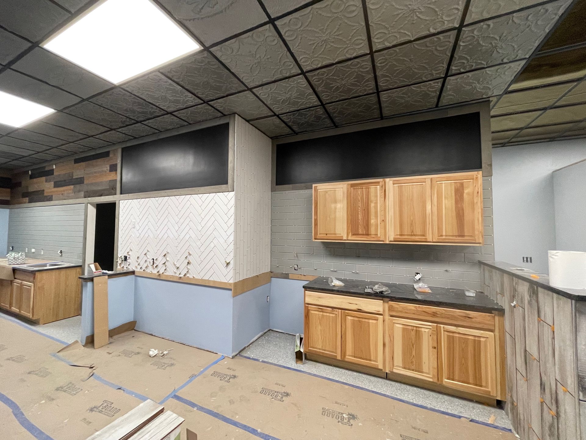 Kitchen under construction with light wood cabinets, black countertops, and grey tile backsplash.