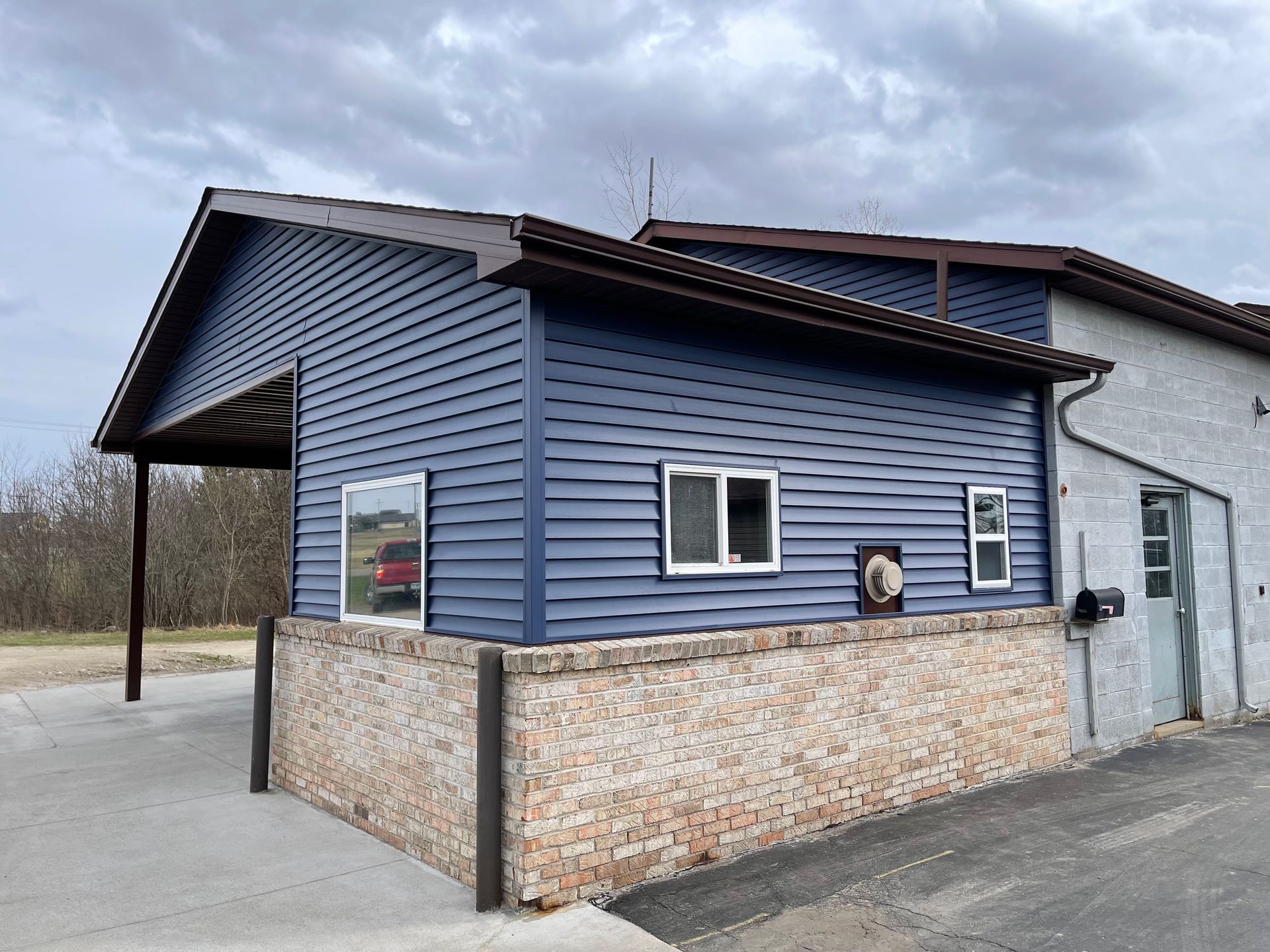 Blue siding building with brown trim, brick facade, and open carport under a cloudy sky.