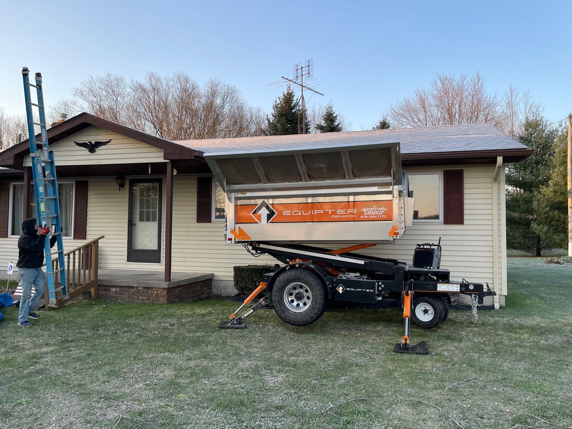 A roofing debris removal trailer next to a house with a ladder propped up.