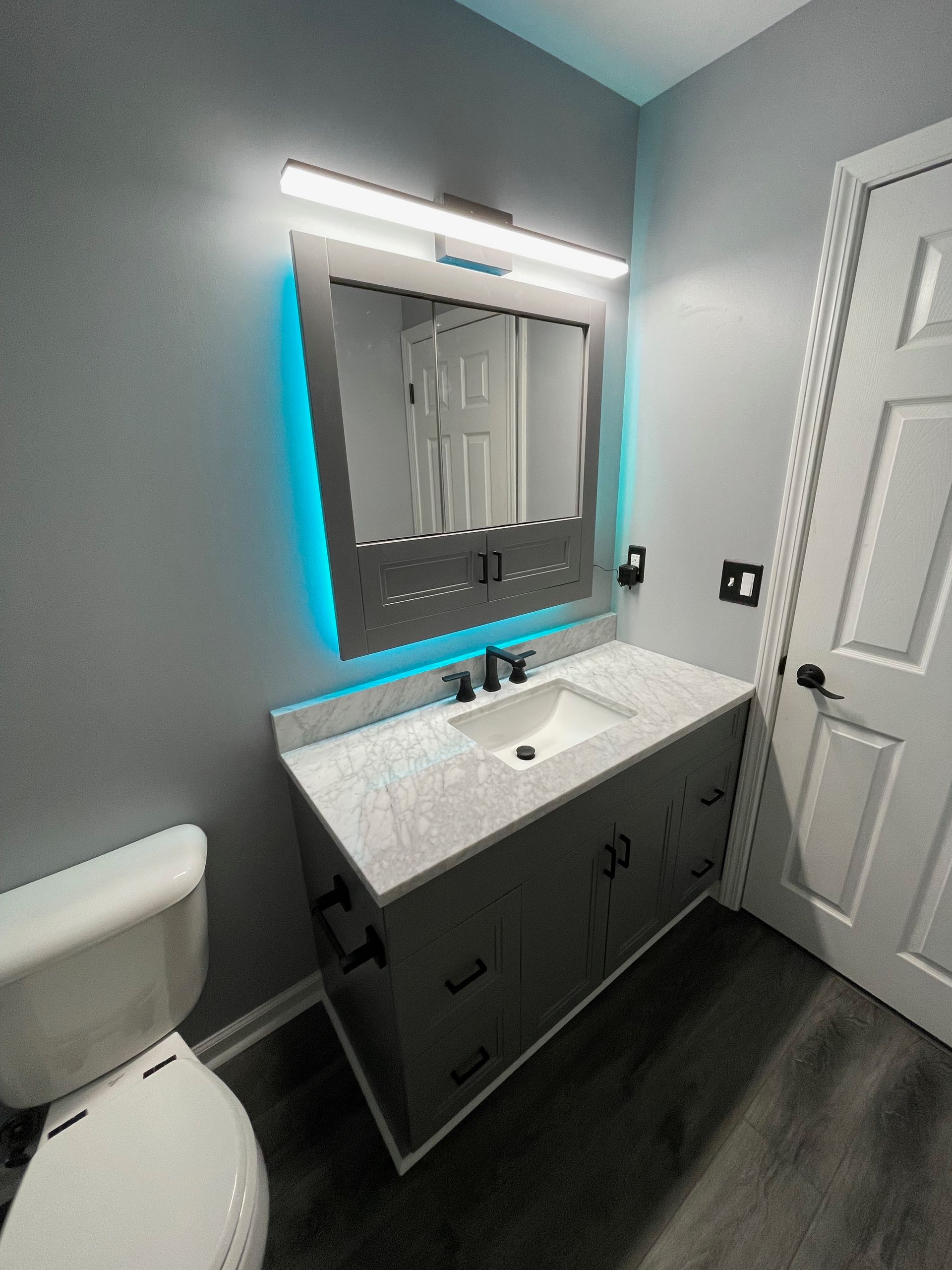 Modern bathroom with gray vanity, countertop, mirror with blue lighting, toilet, and door.