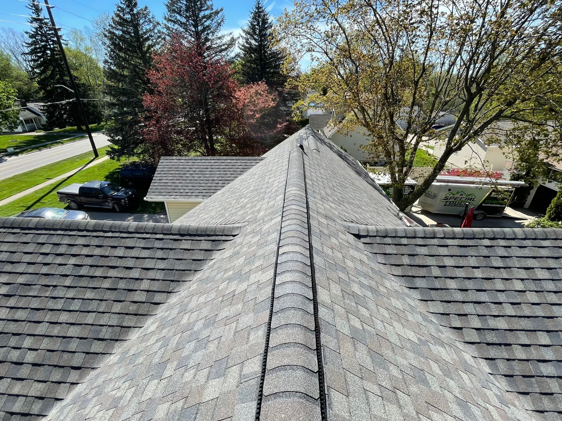 View of a newly shingled roof with a central ridge, surrounded by trees and residential properties.