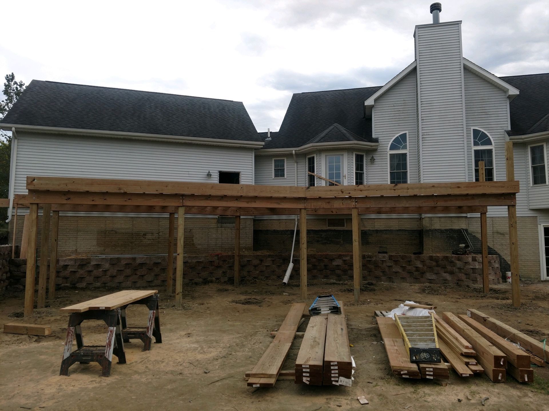 Deck under construction behind a two-story house. Lumber, tools, and a partial frame are visible.