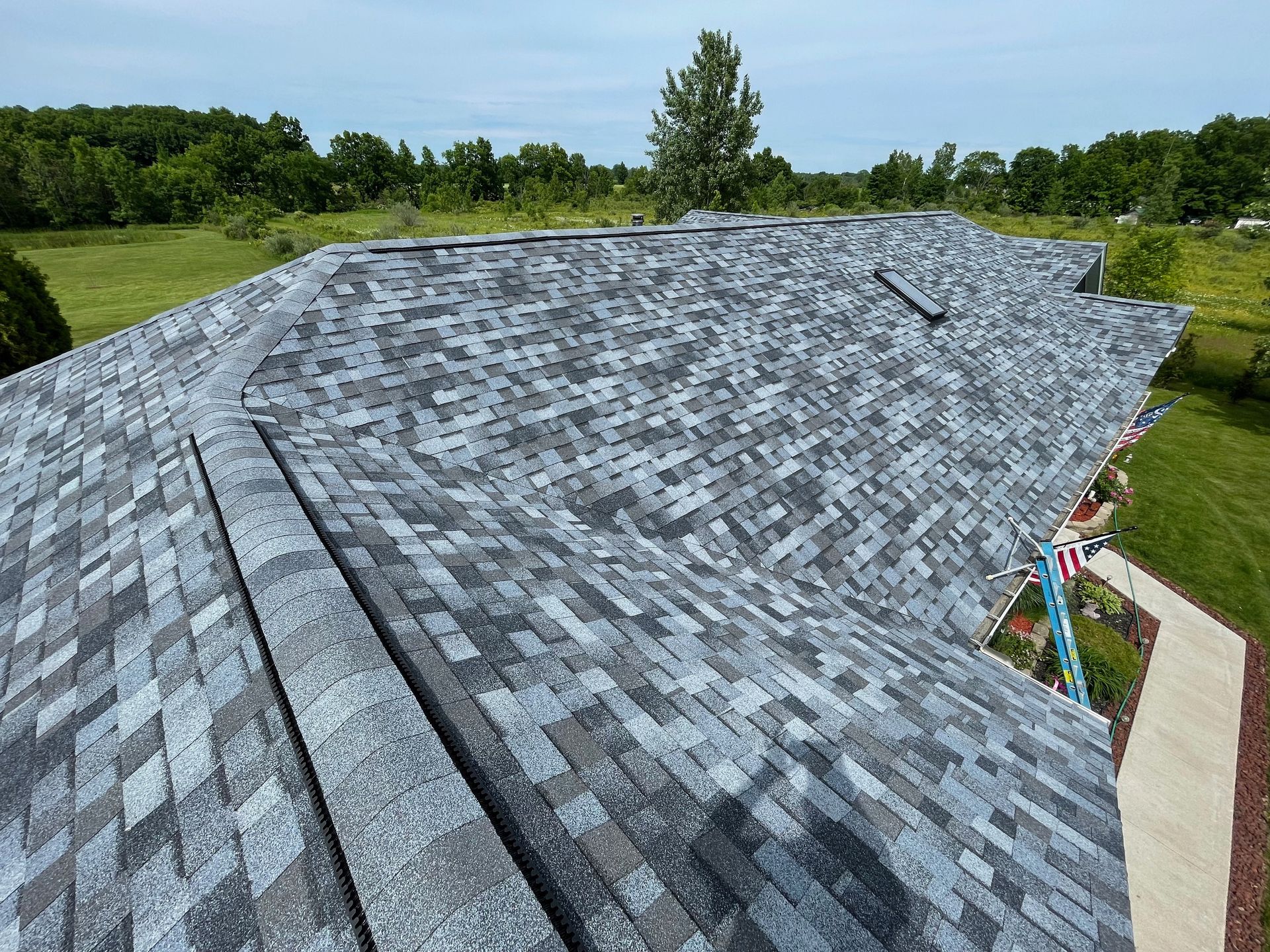 Overhead view of a house roof with new gray asphalt shingles. Green trees and sky in the background.