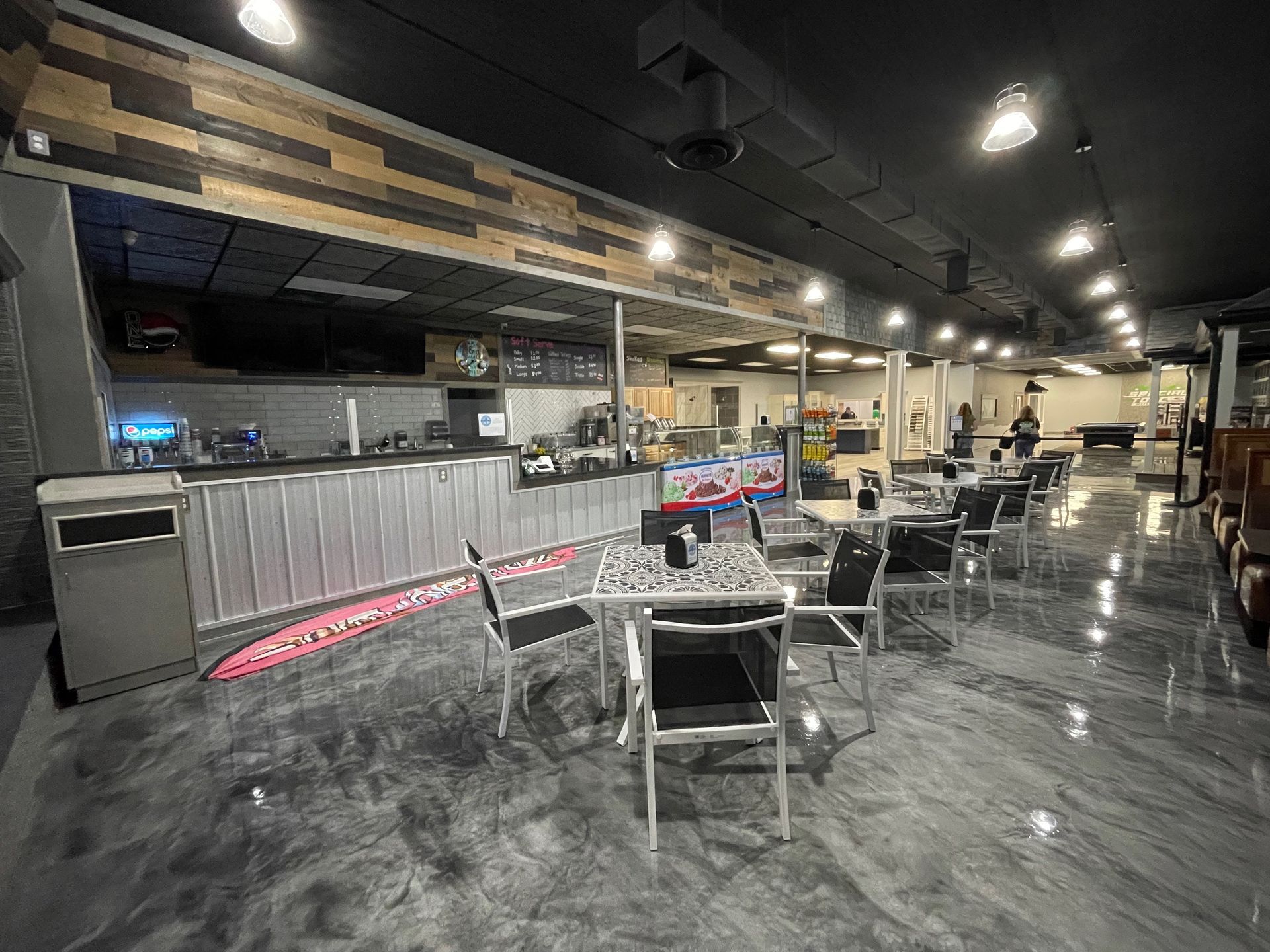 Bar and seating area; metal bar, tables with chairs, and glossy floor in dimly lit room.