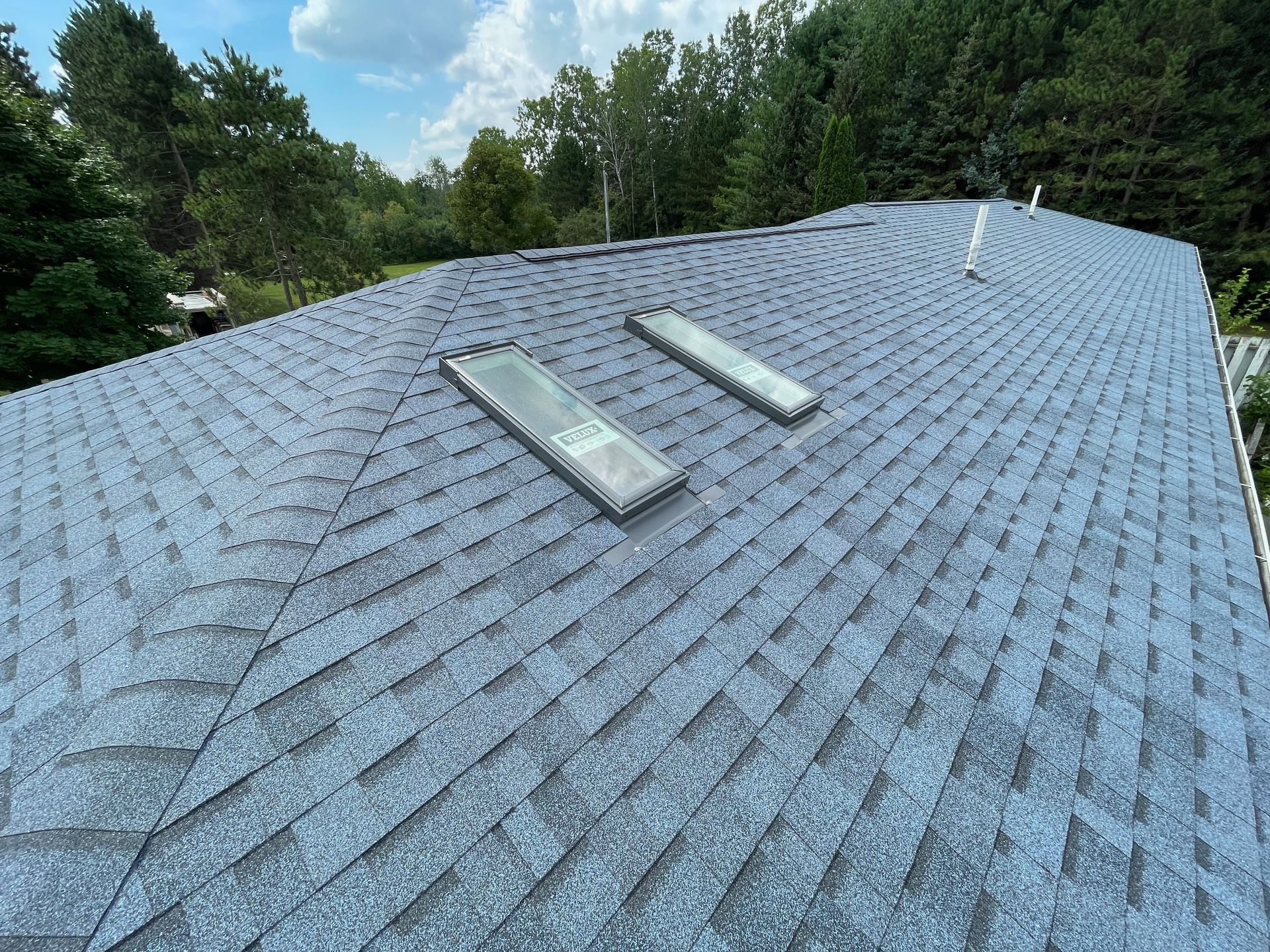 View of a blue-gray shingle roof with two rectangular skylights, surrounded by trees under a partly cloudy sky.