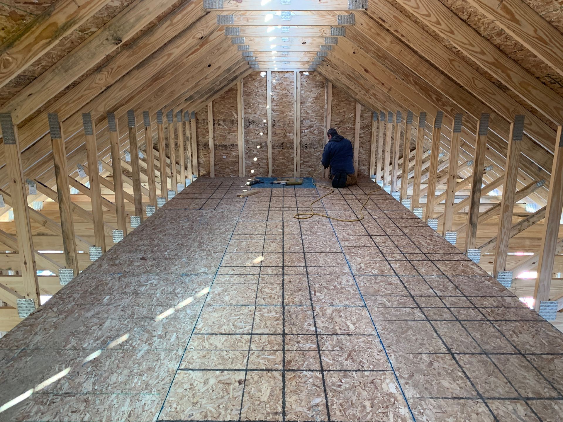 Interior view of an unfinished attic with plywood flooring, wooden rafters, and a person working in the distance.