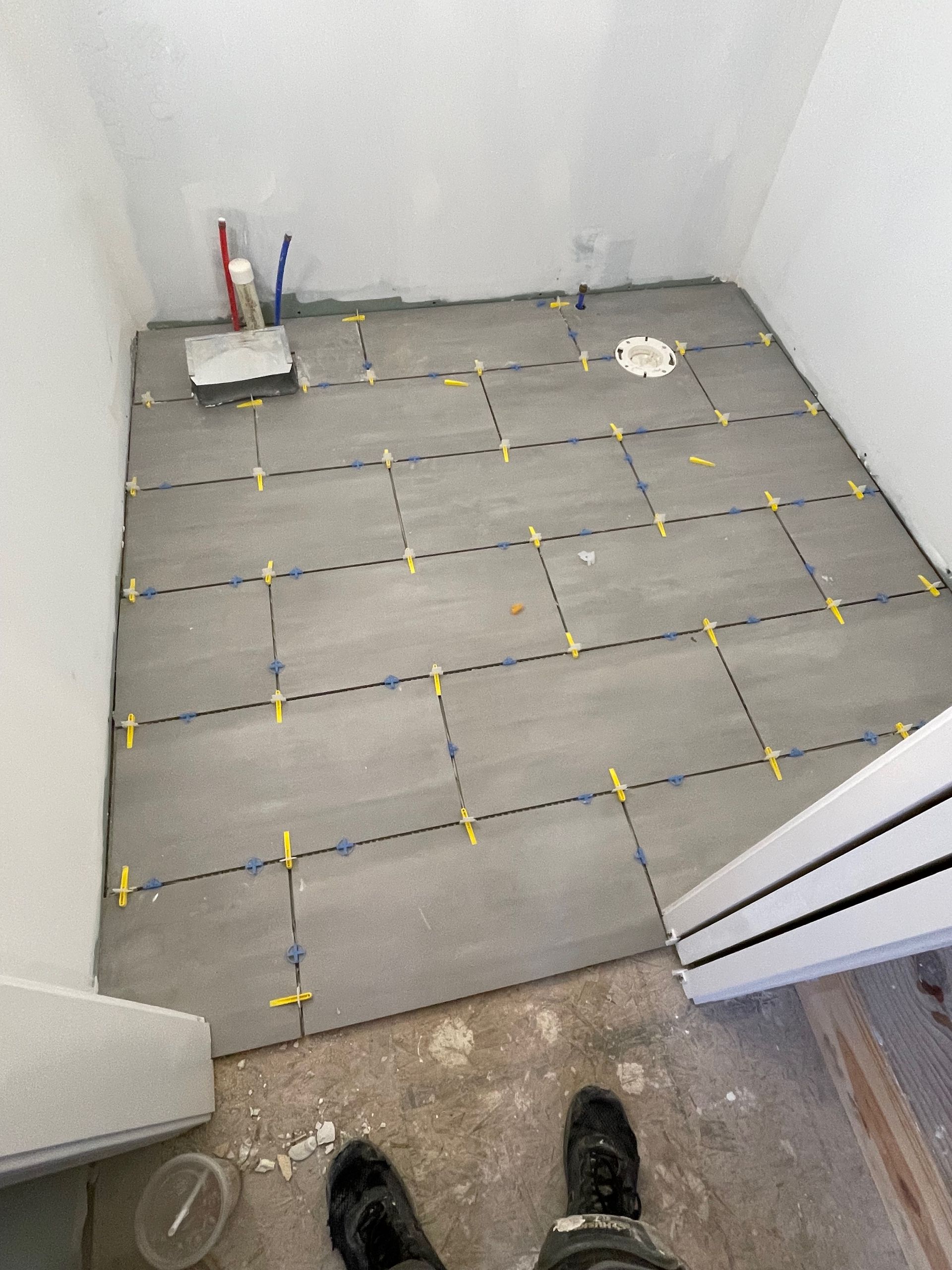 Bathroom floor with gray tiles laid out, spacers in place, near plumbing and door frame.