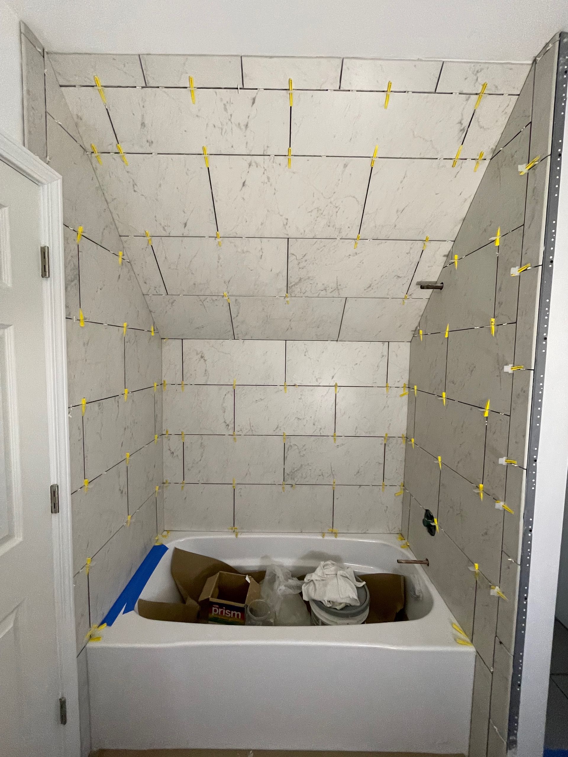 Bathroom tub alcove tiled with light-colored rectangular tiles; spacers used; blue tape on tub.