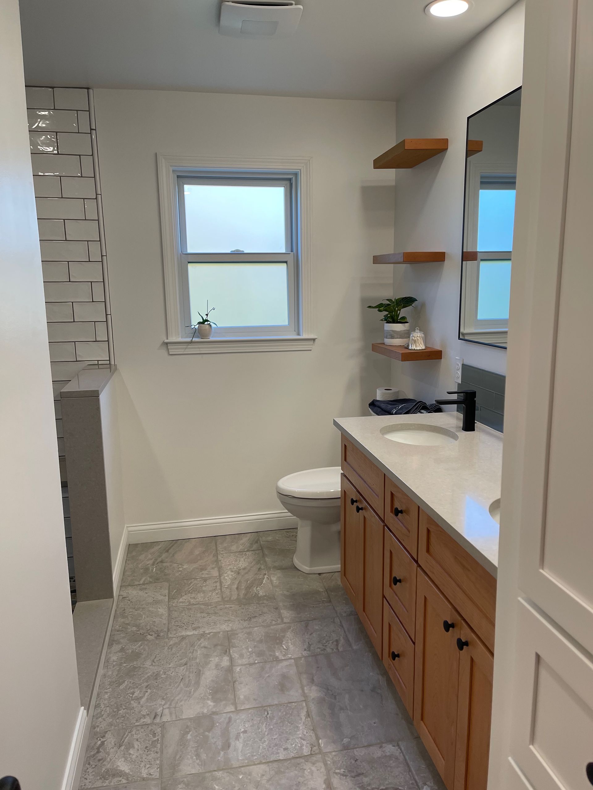 Bathroom with wood vanity, floating shelves, and gray tile flooring. White walls and a window.