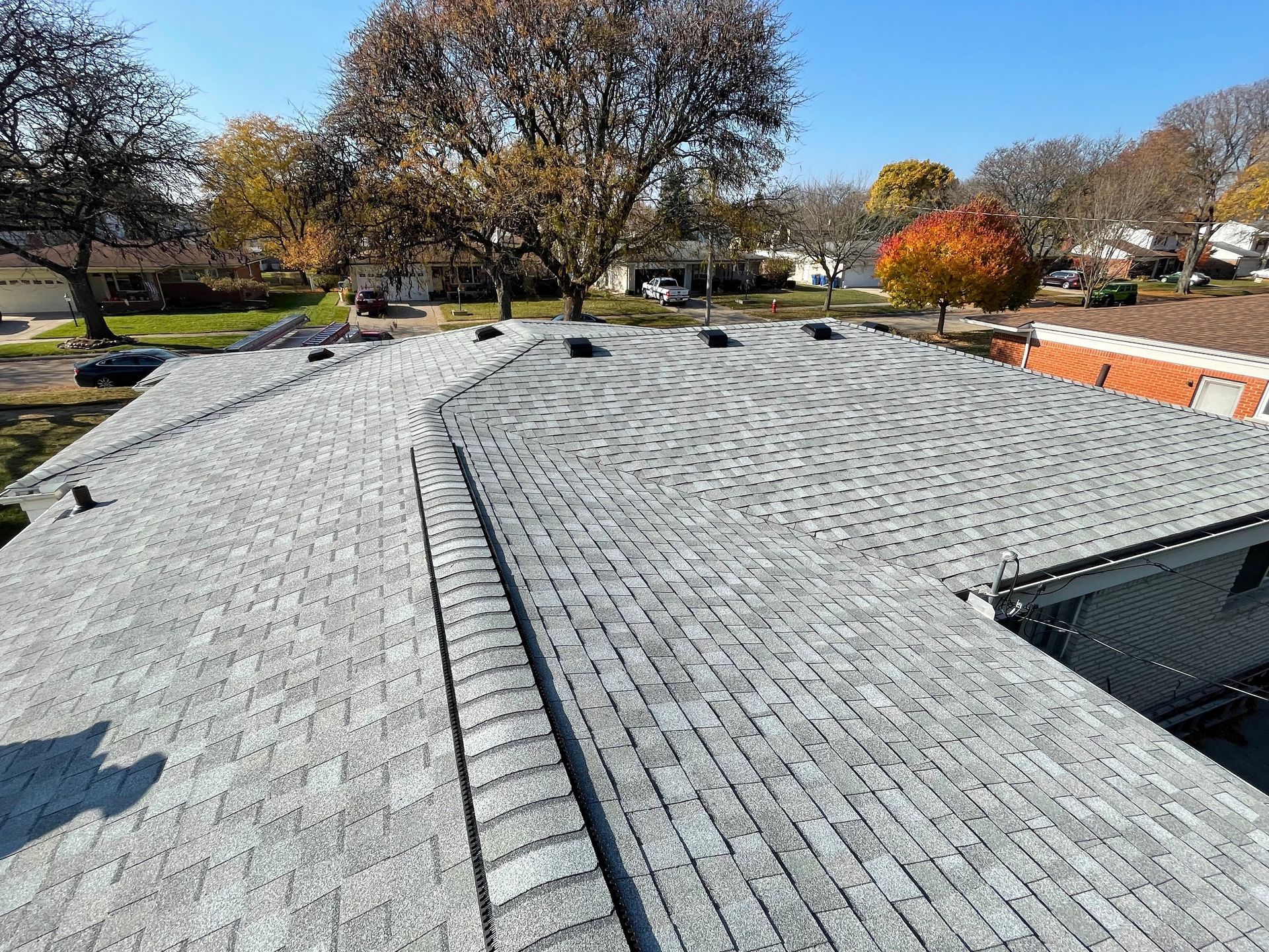Gray asphalt shingle roof on a house, view from above, clear blue sky, trees in the background.