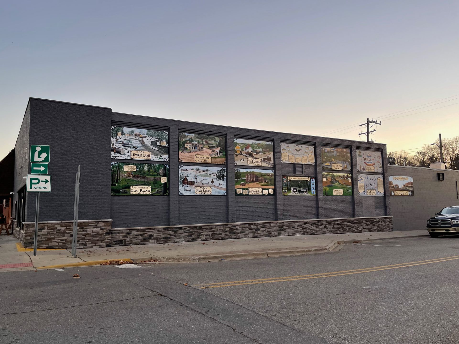 Building with a mural depicting multiple scenes; street, parking sign visible.