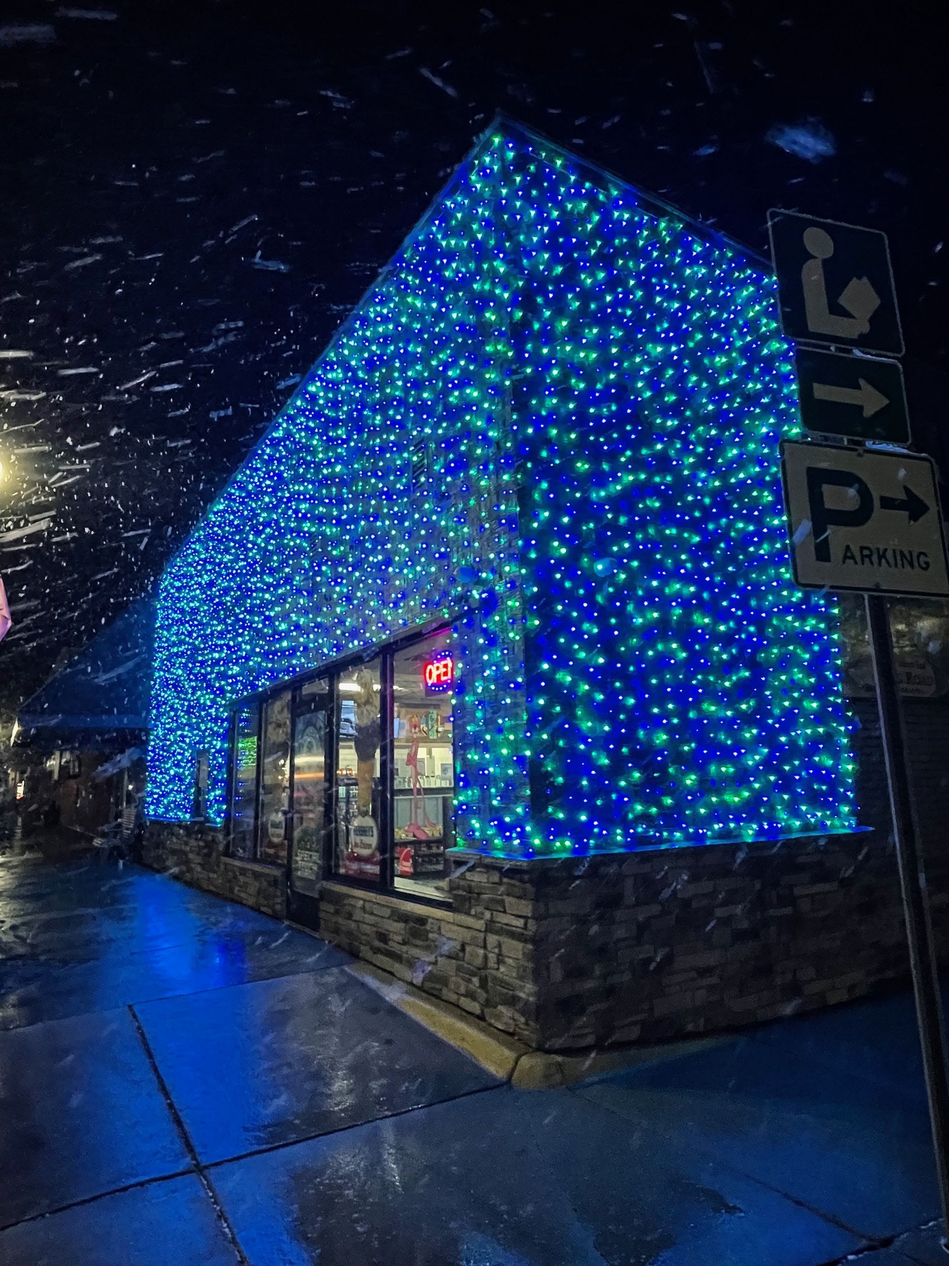 A storefront covered in blue and green string lights, illuminated at night. Snow is falling.