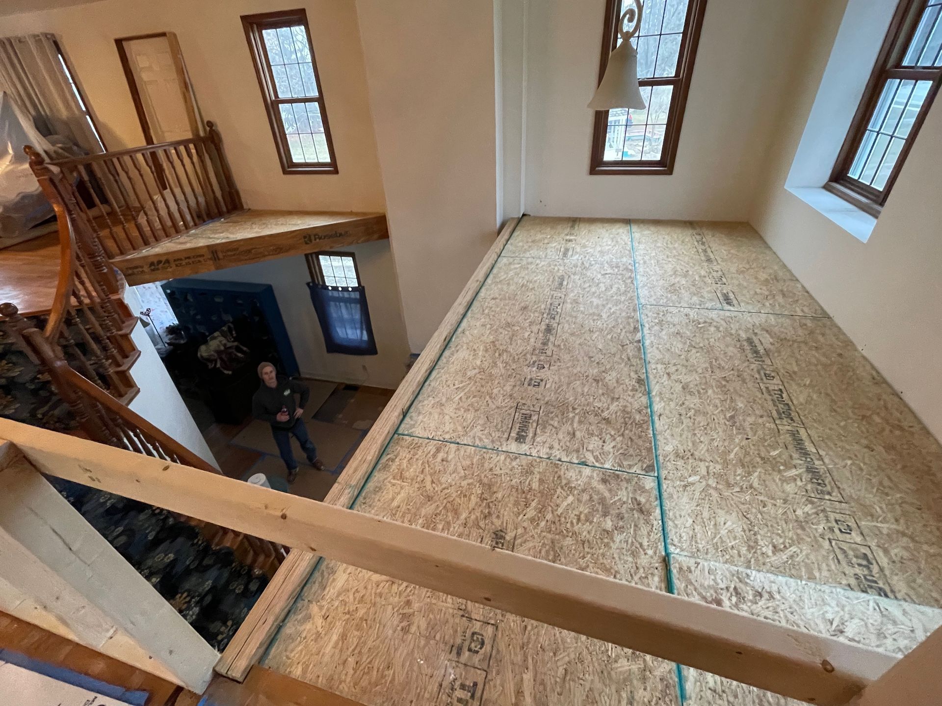 Interior view: unfinished loft with OSB flooring, wooden handrail, windows, person standing below.