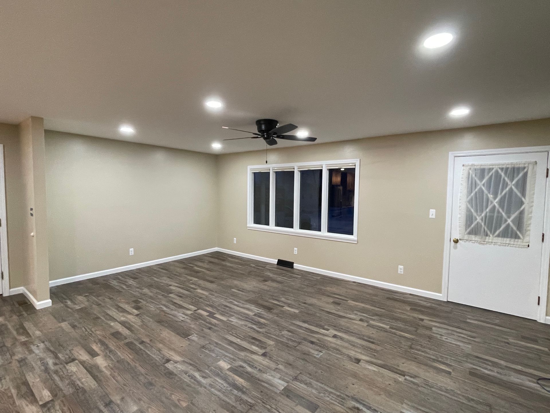 Empty room with beige walls, dark wood-look flooring, large window, and white door.