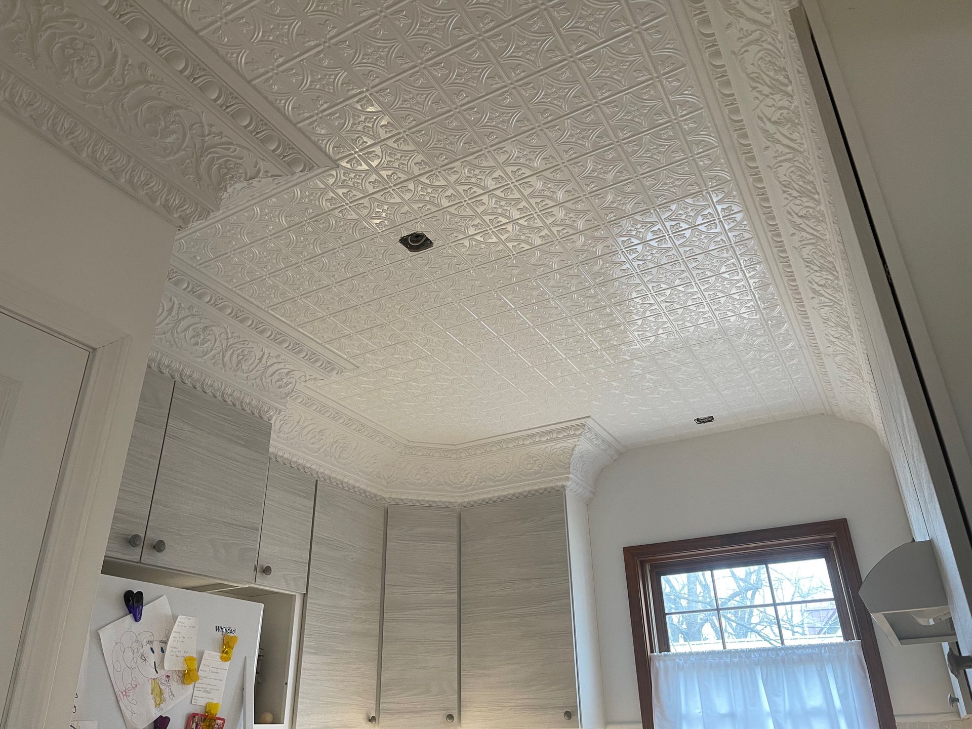 White ornate tin ceiling in a kitchen with cabinets, a window, and a light fixture.