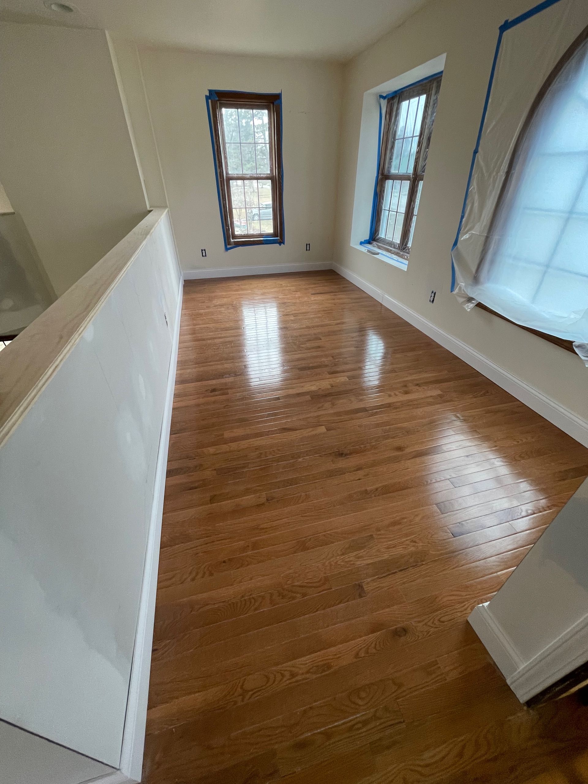 Newly finished hardwood floor in a room with two windows and a white railing.