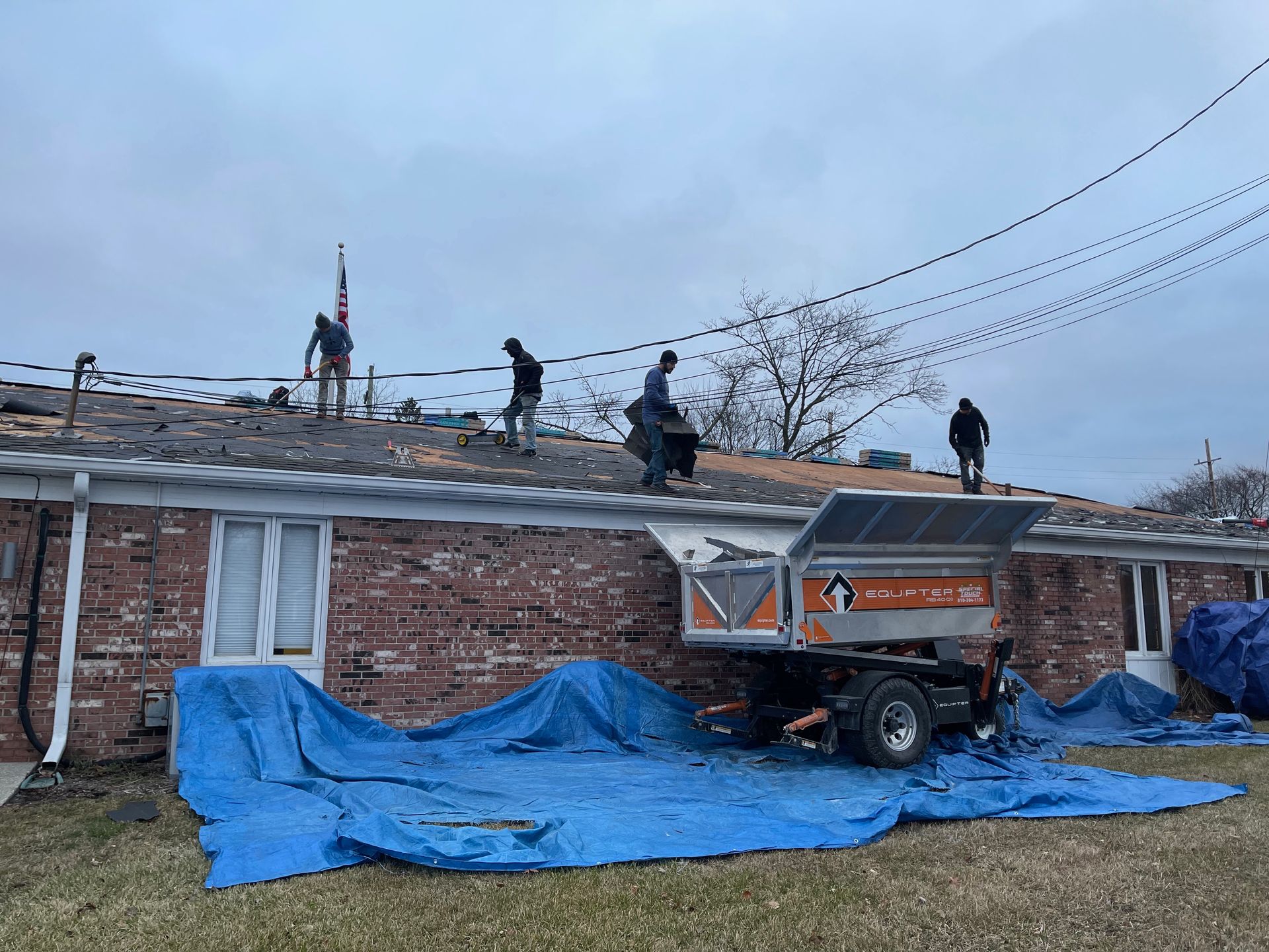 Workers repairing a roof, some carrying debris. Blue tarp covers the lawn; cloudy sky.