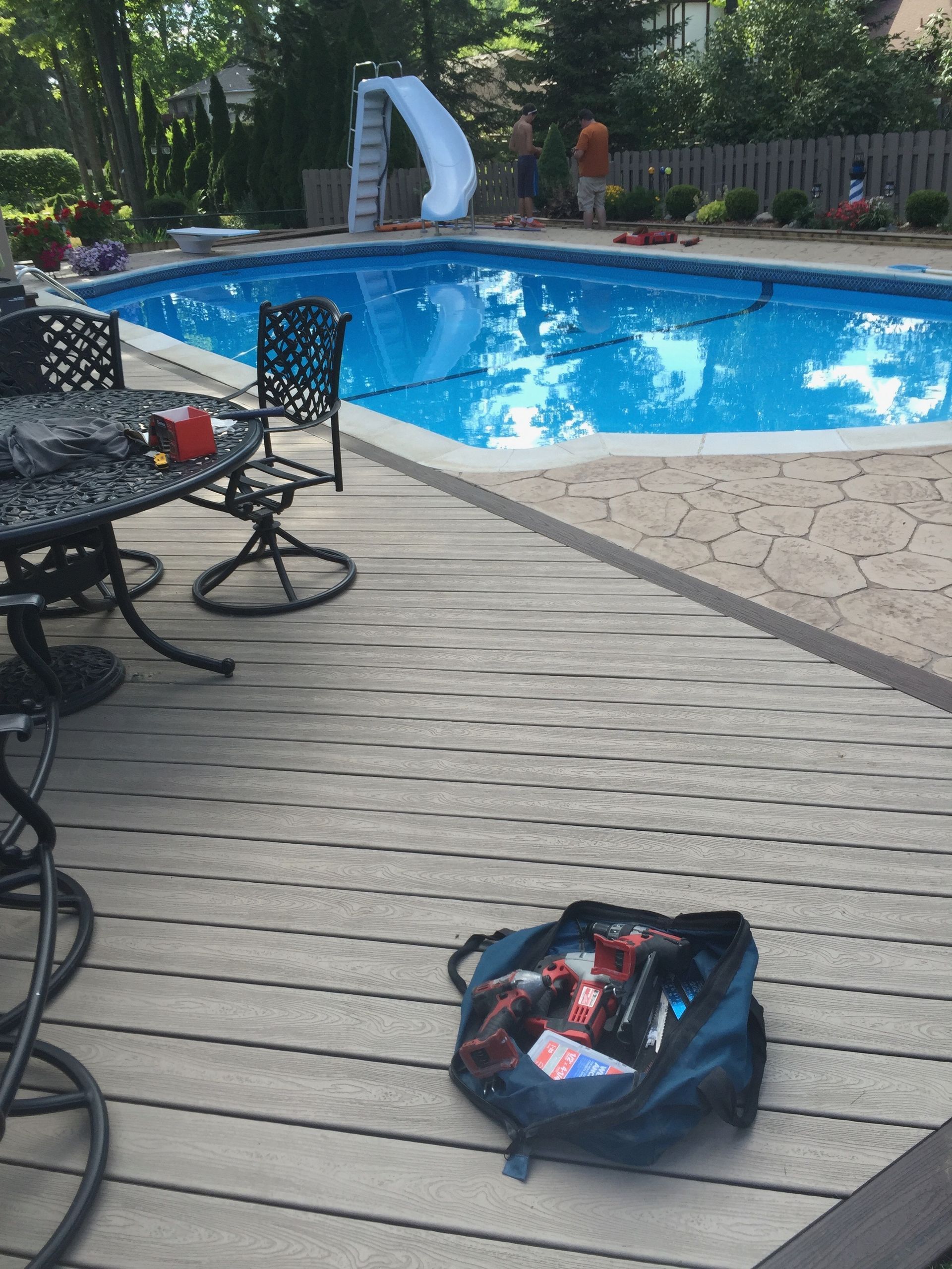Backyard pool with a slide, deck, and a bag. People are in the background. Sunny day.