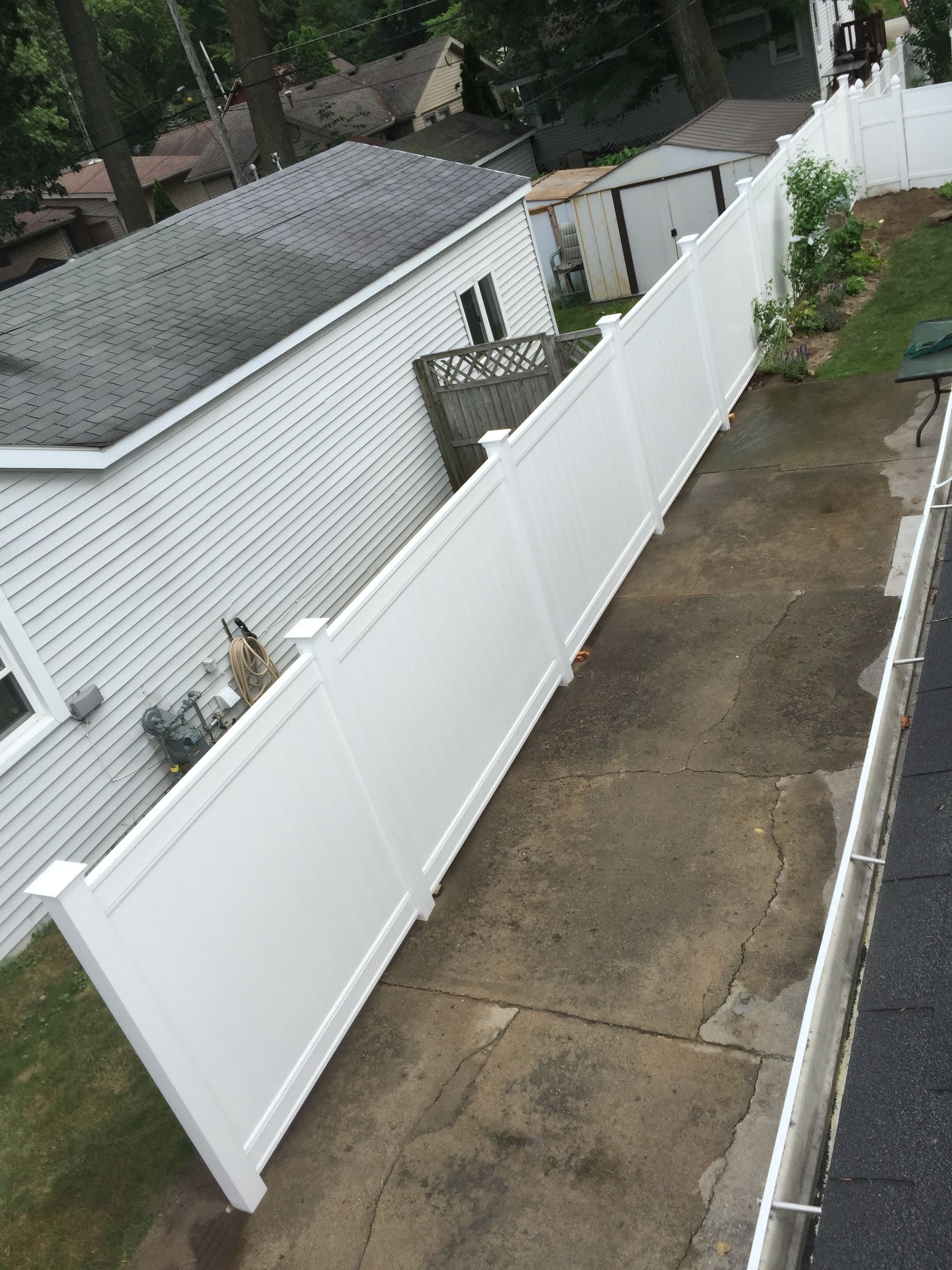 White vinyl fence along a concrete driveway beside a white building and small shed.