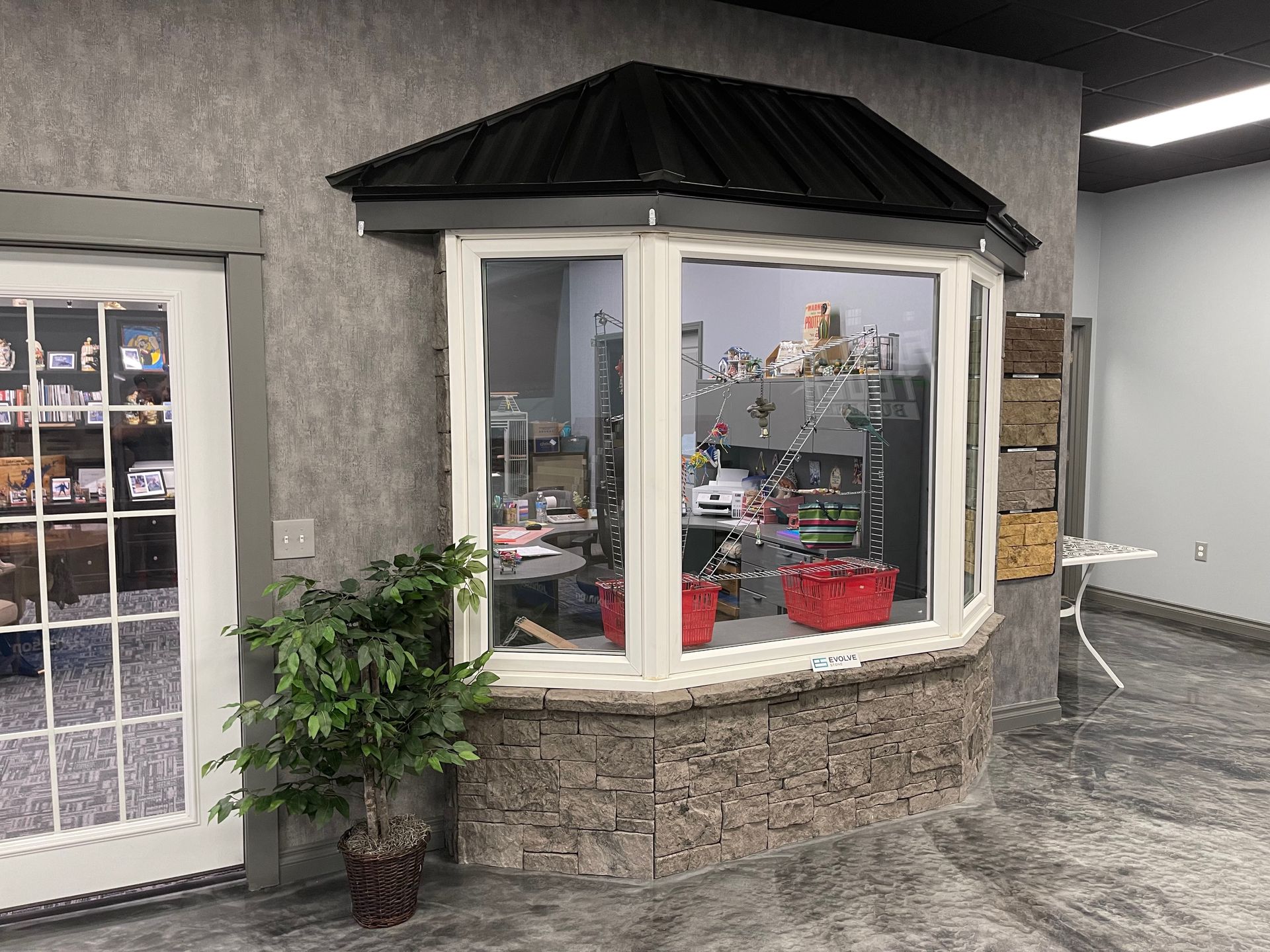 Bay window with black roof, faux stone facade, and display items visible inside. A door and small plant are to the left.