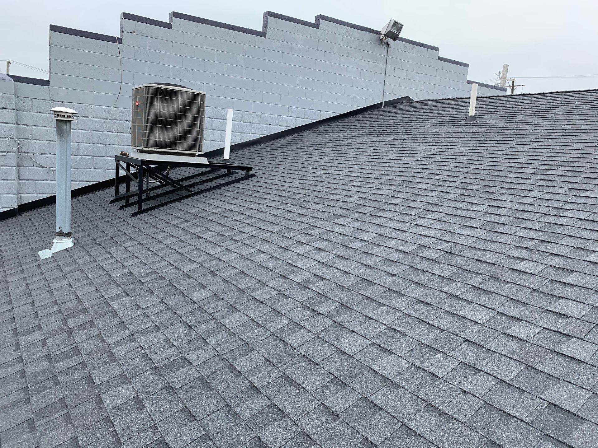 Grey asphalt shingle roof with an AC unit and vent pipes, against a light grey building exterior.