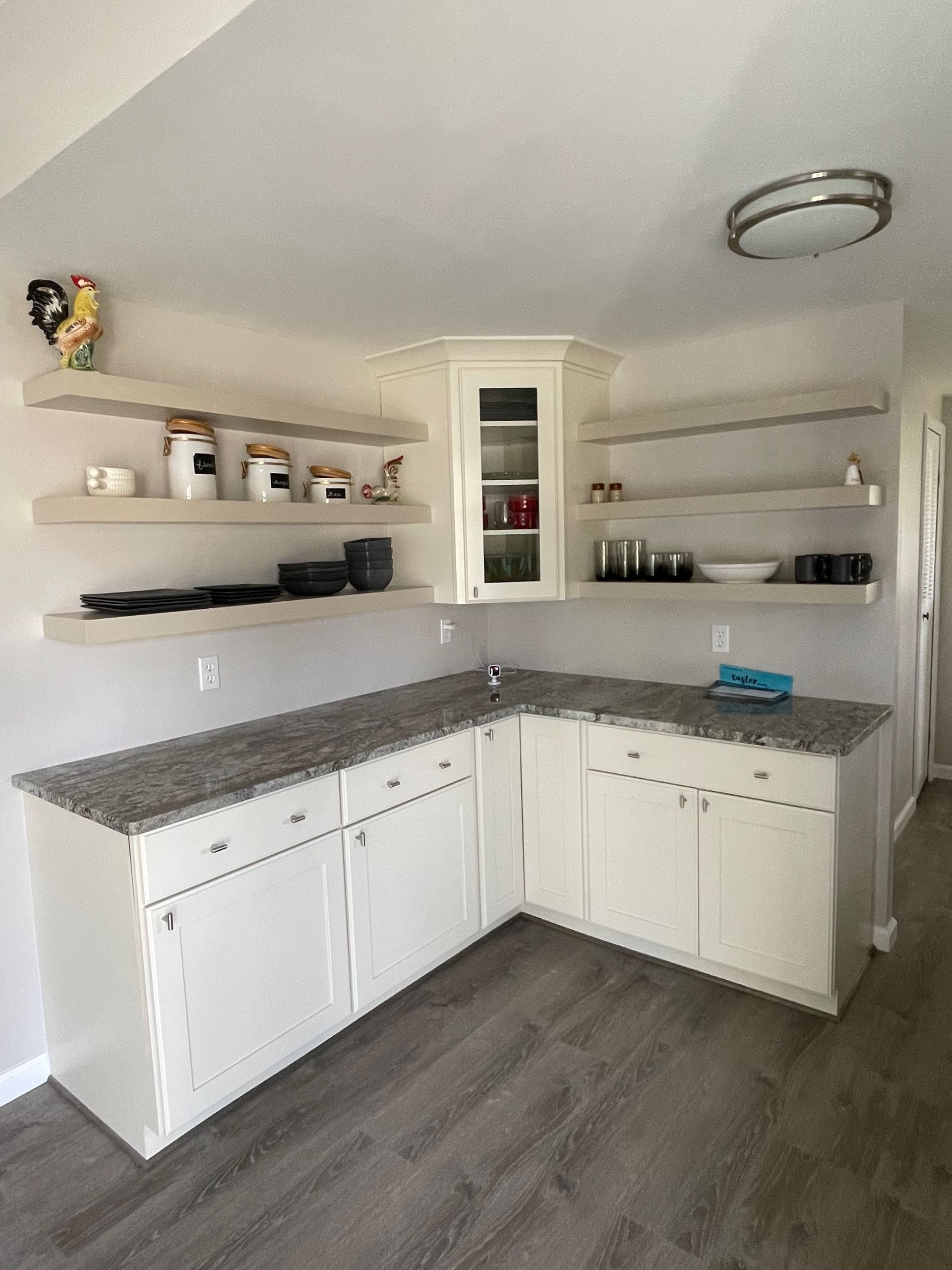 L-shaped kitchen with white cabinets, gray countertops, floating shelves, and a glass-door cabinet.