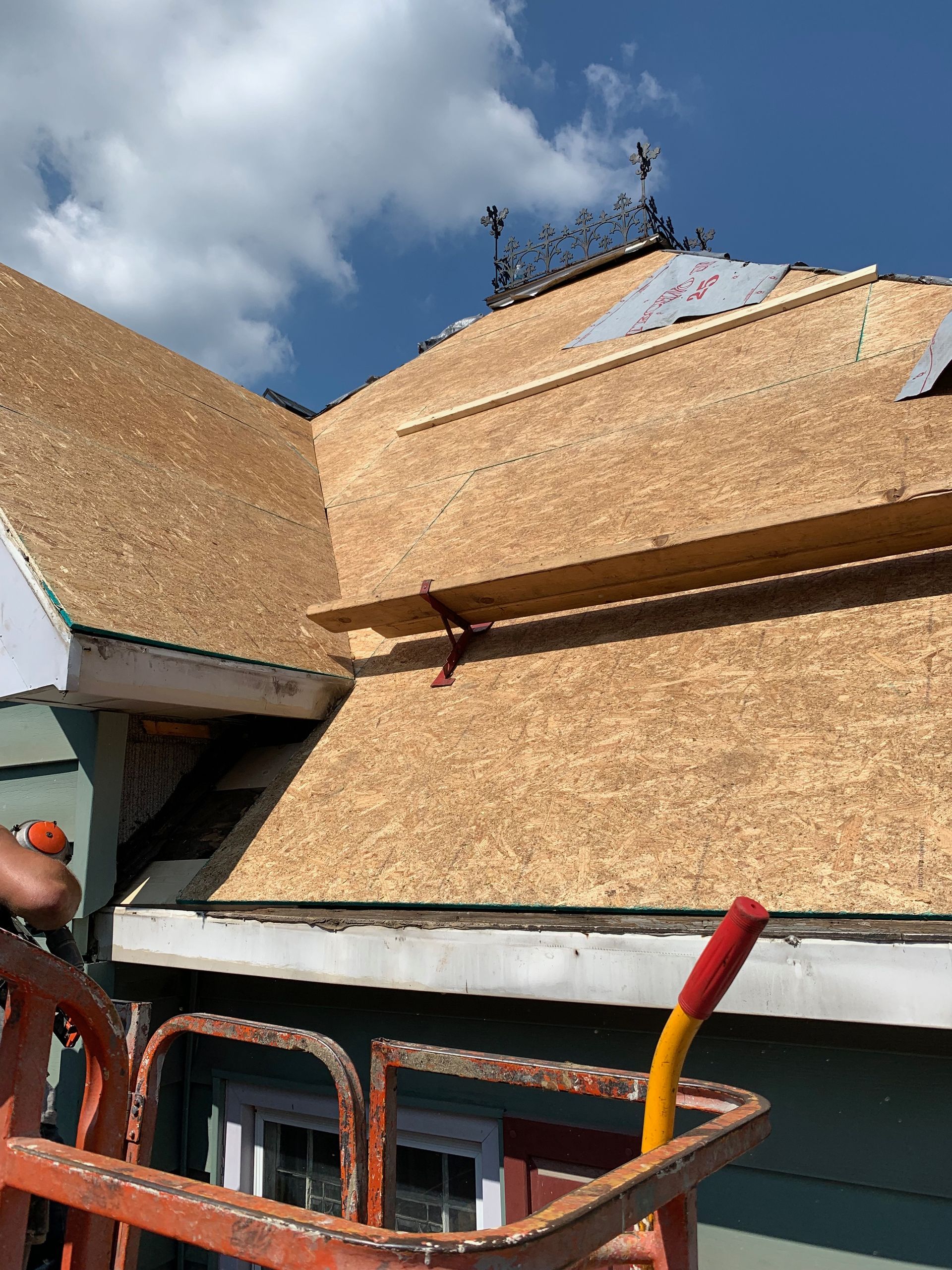 Roof of a building under construction, showing plywood and roof material, with a lift in the foreground.