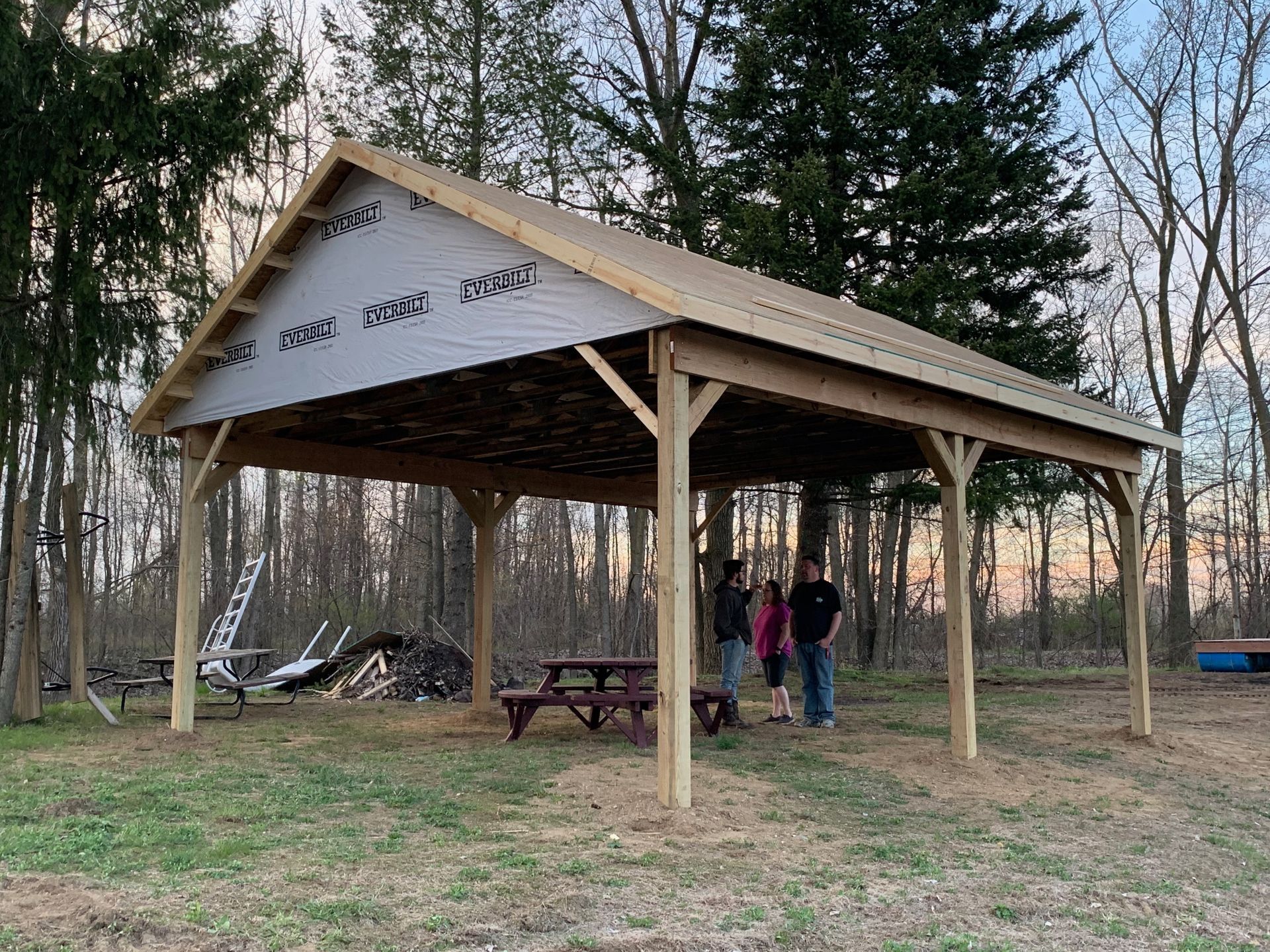 Wooden pavilion with people standing under it; picnic table, trees.