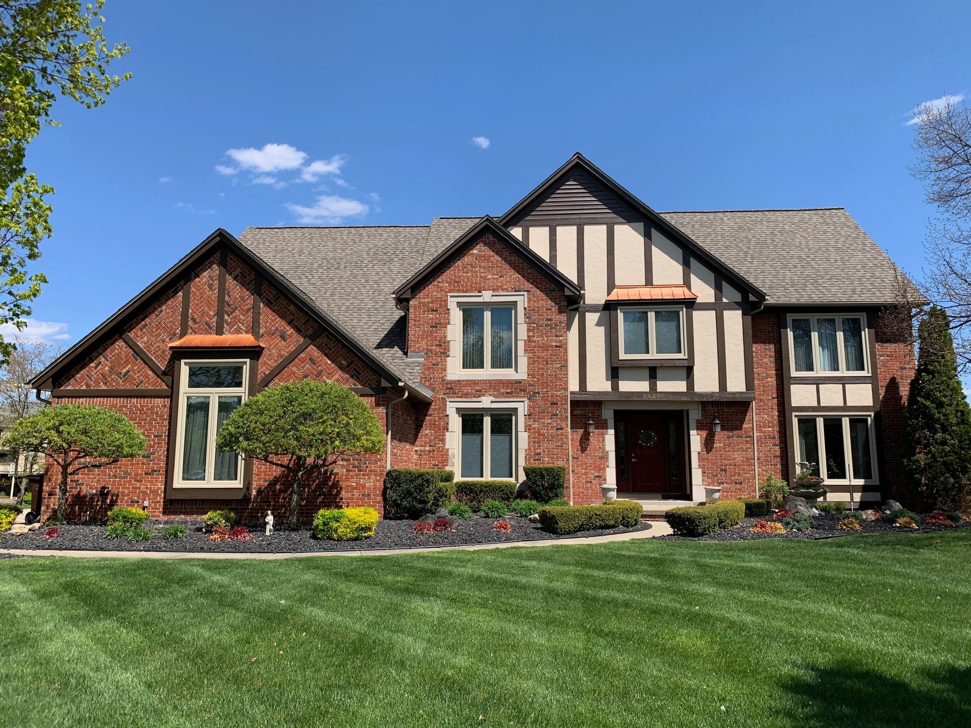 Two-story brick Tudor-style house with green lawn, flower beds, and a blue sky.