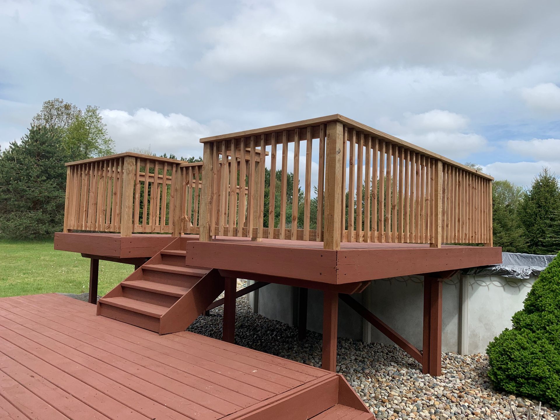 Wooden deck with stairs, built around an above-ground pool, brown and red tones, with surrounding green lawn and cloudy sky.