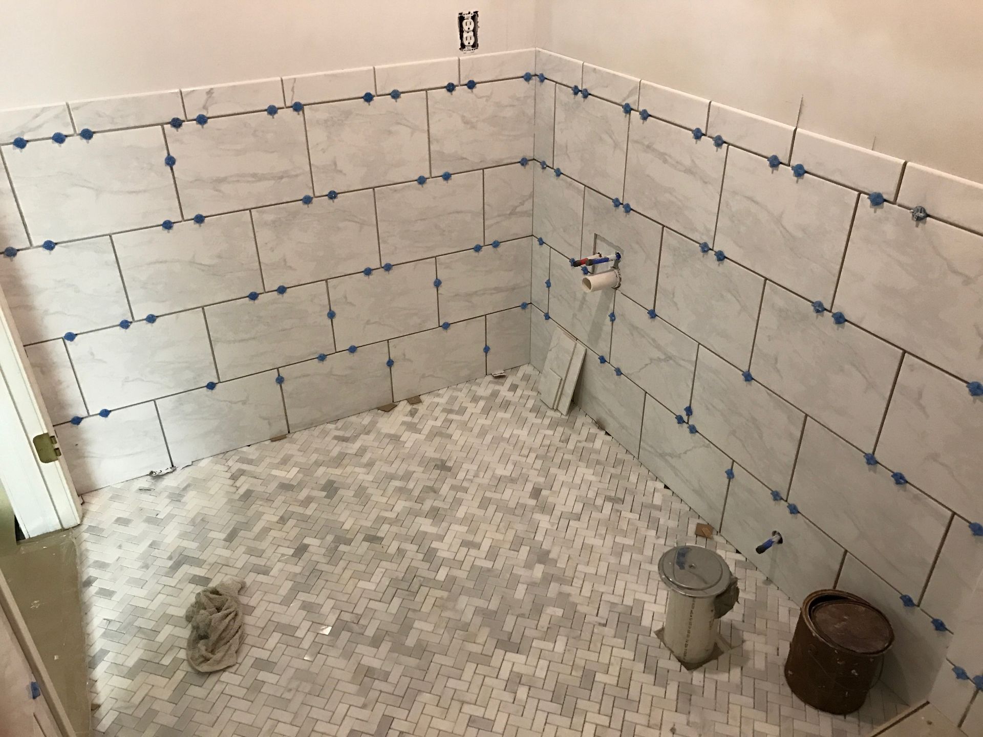 Bathroom under construction, walls tiled in white marble, small mosaic floor tiles, buckets and tools present.