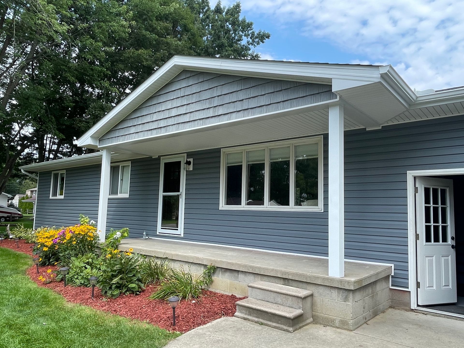 Blue-gray house with white trim, porch, and windows. Landscaping in front. Cloudy sky.